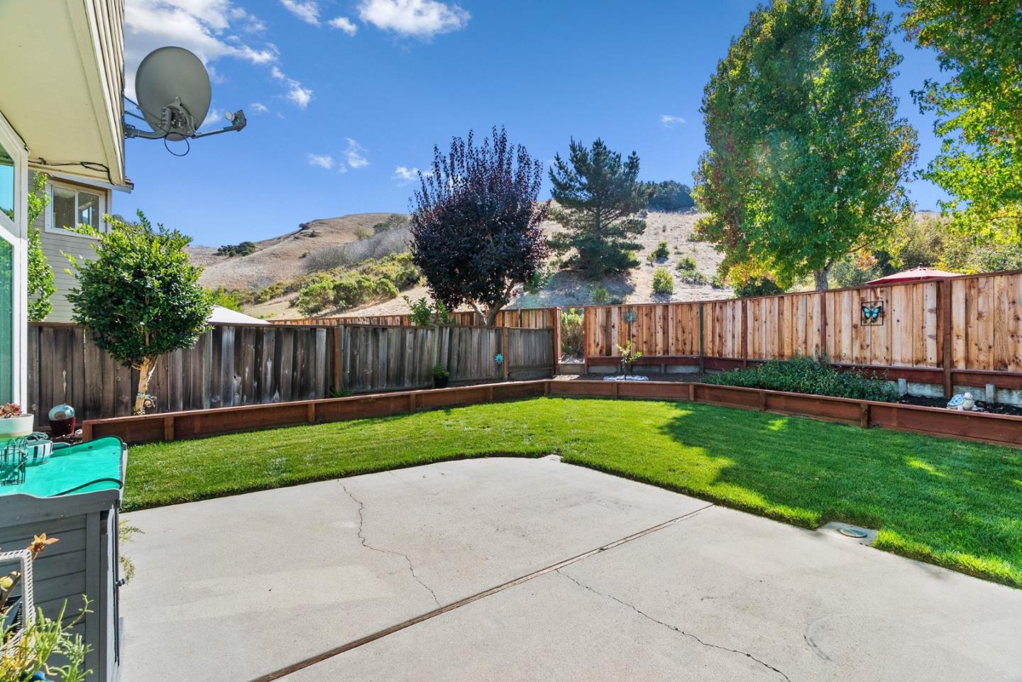 Fenced backyard with a concrete patio, green lawn, raised planting beds, and rolling hills beyond.