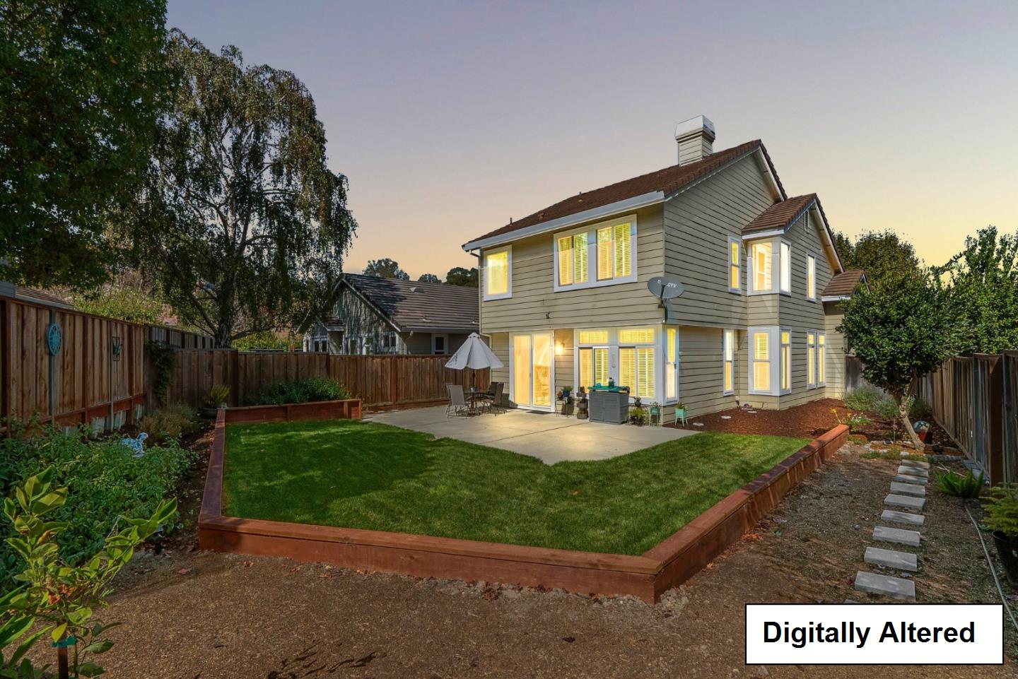 Two-story house at dusk with a raised backyard lawn, fenced patio, and illuminated windows.