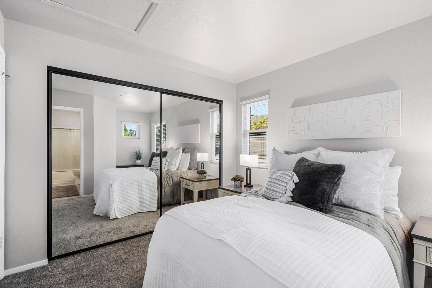 Staged bedroom with a white-and-gray bed, mirrored closet doors, and gray carpet.
