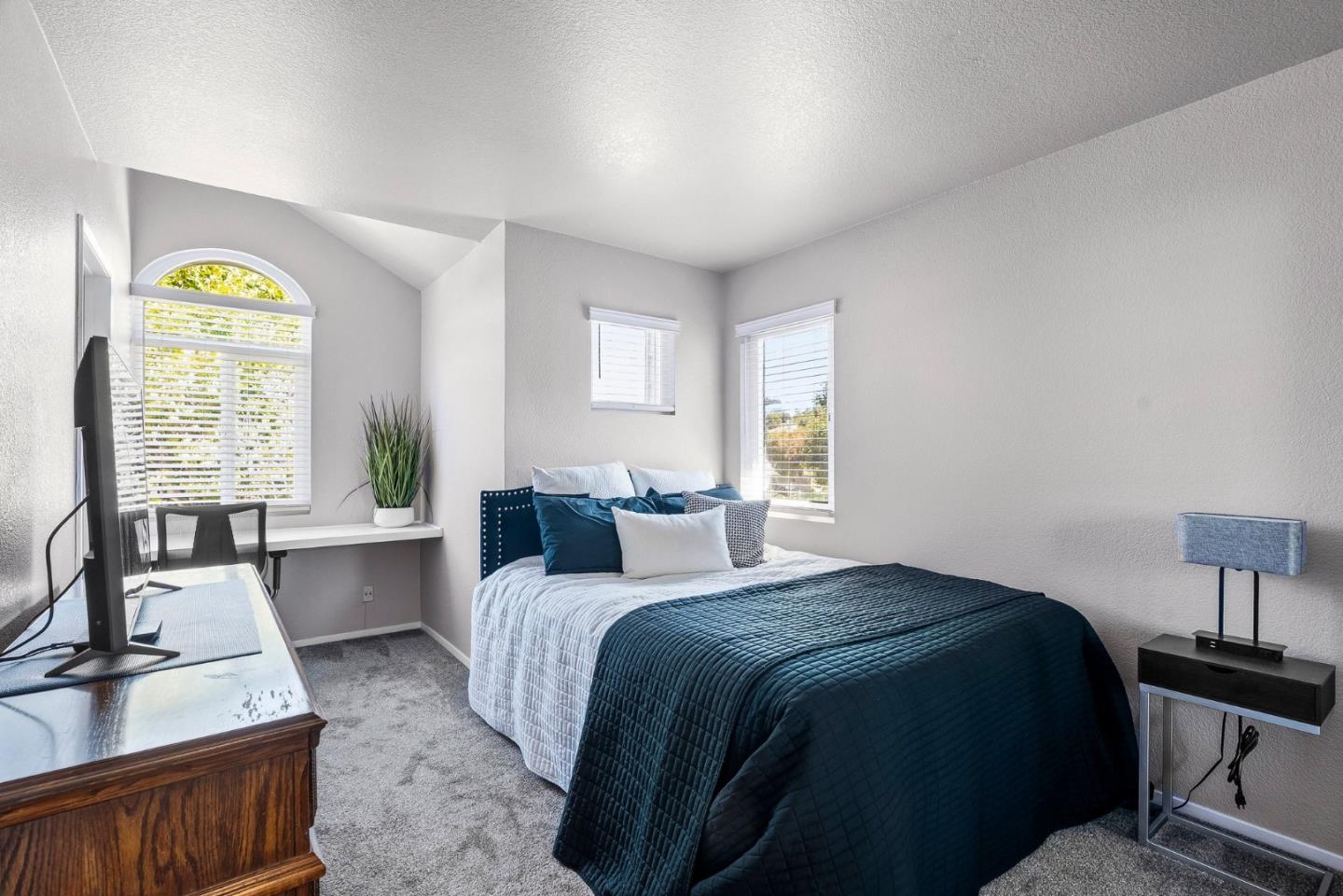 Bedroom with dark teal bedding, gray carpet, and a desk beneath an arched window.