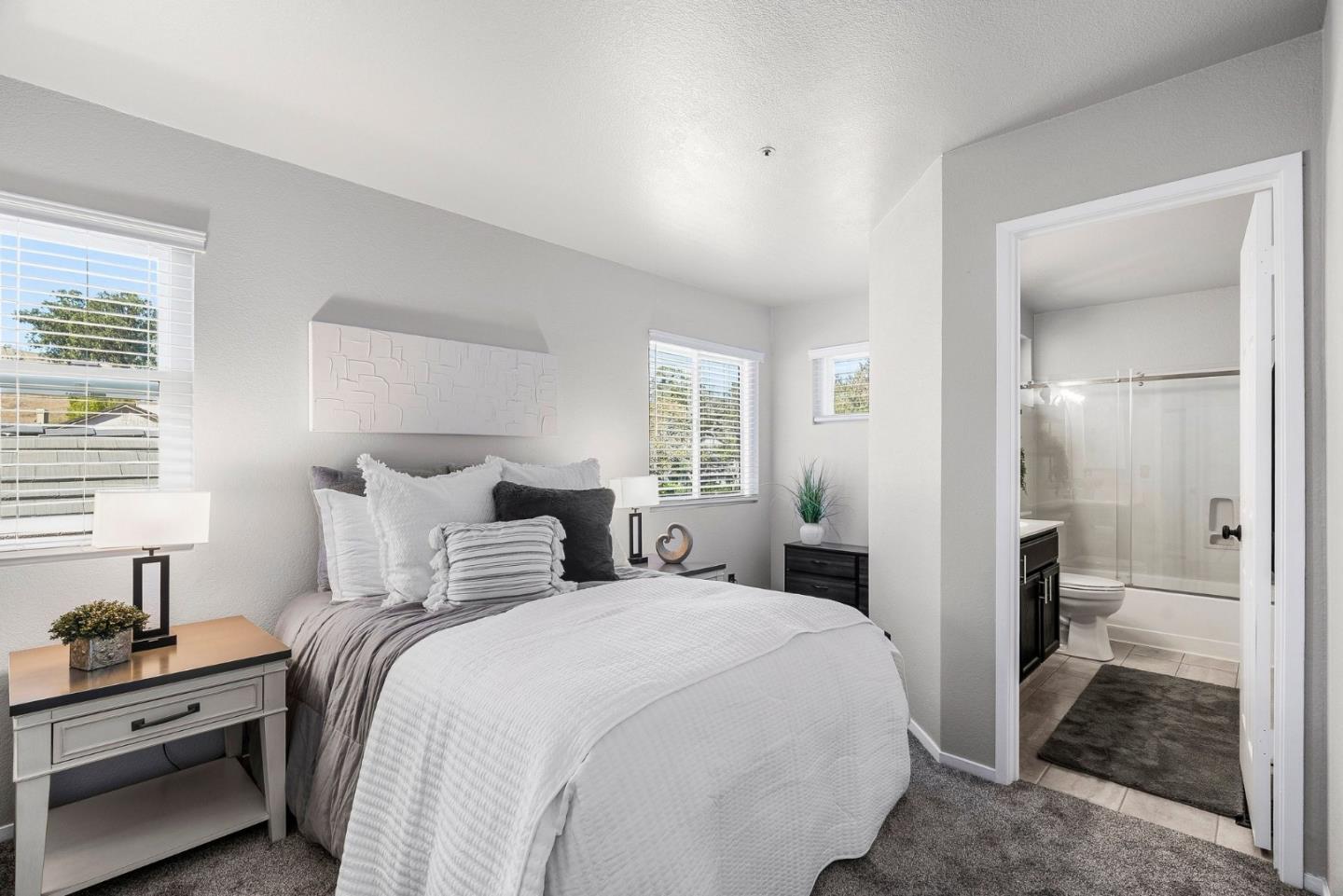 Staged bedroom with white and gray bedding and an open doorway to an adjoining bathroom.