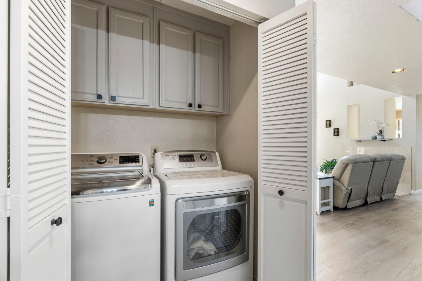 Laundry closet with washer and dryer beneath cabinets, opening toward a bright living room.