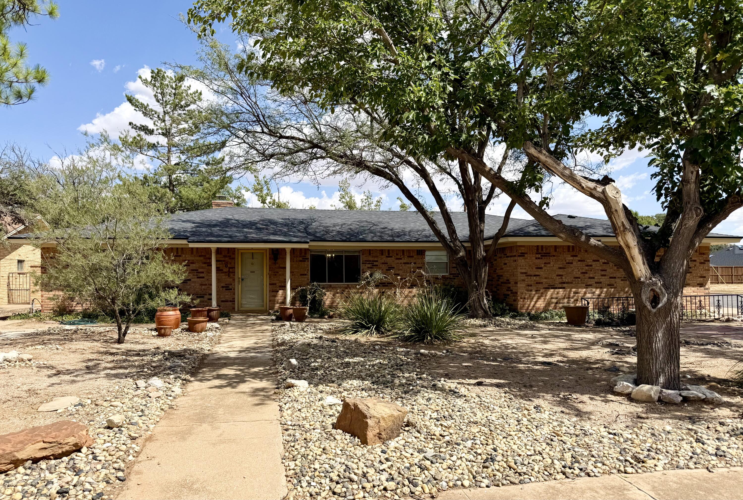 Single-story brick house shaded by mature trees, with a rock garden and walkway to the front door.