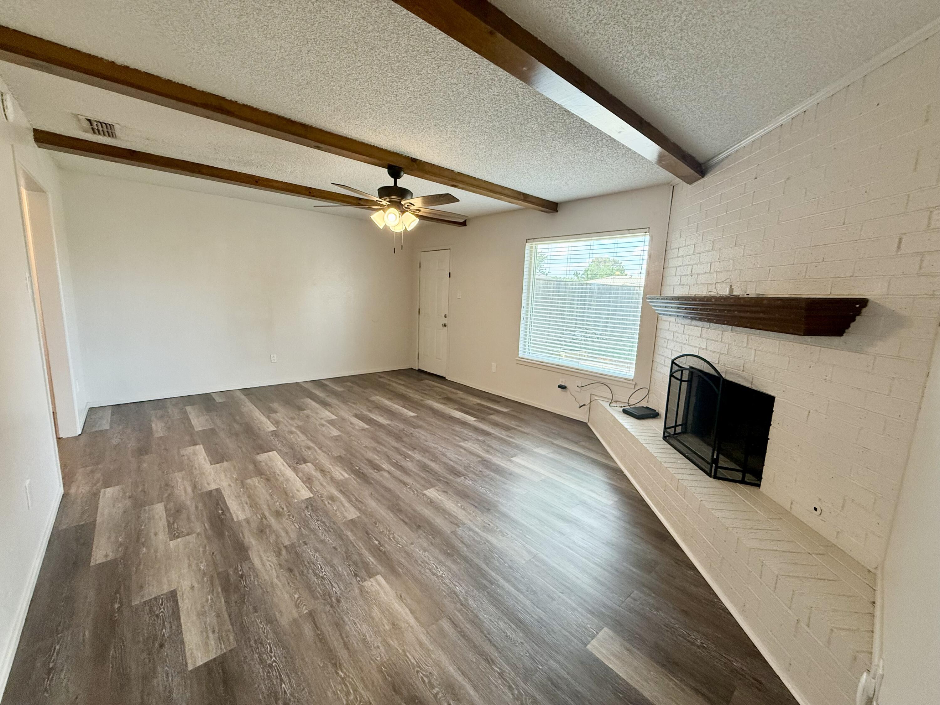 Empty living room with exposed wood ceiling beams, a brick fireplace, and gray-brown plank flooring.