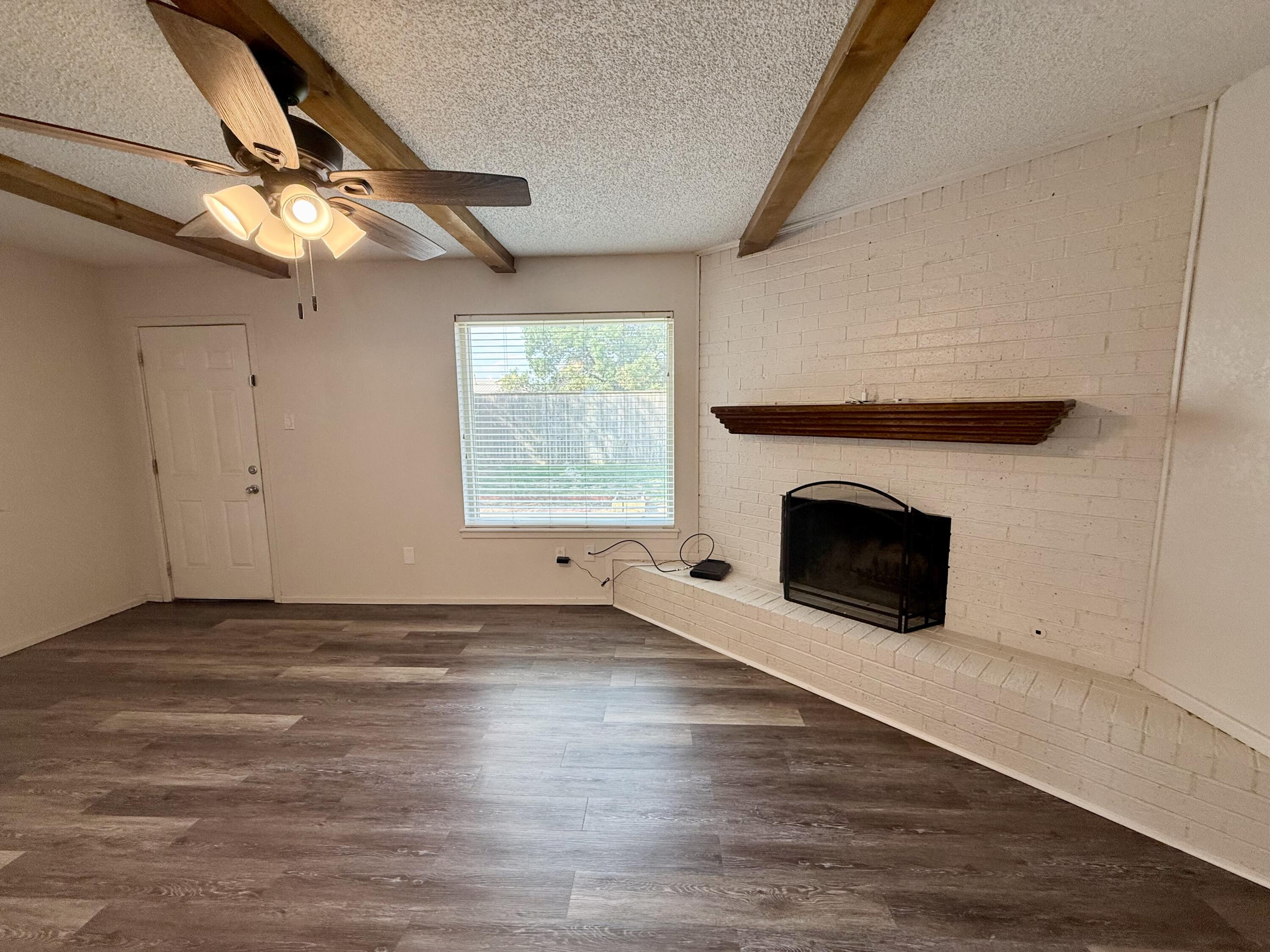 Empty living room with exposed beams, dark flooring, and a white brick fireplace.