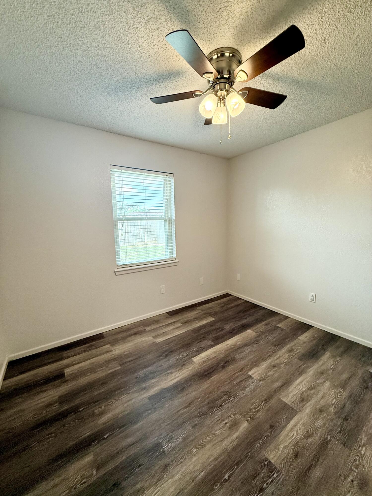 Empty room with dark wood-look flooring, a window with blinds, and a lit ceiling fan.
