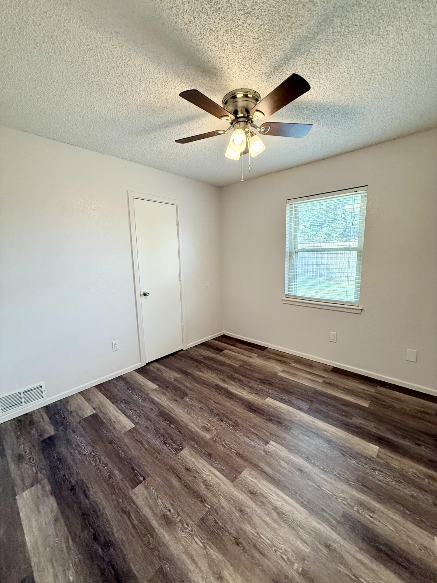 Empty room with dark plank flooring, a lit ceiling fan, and a window with blinds.