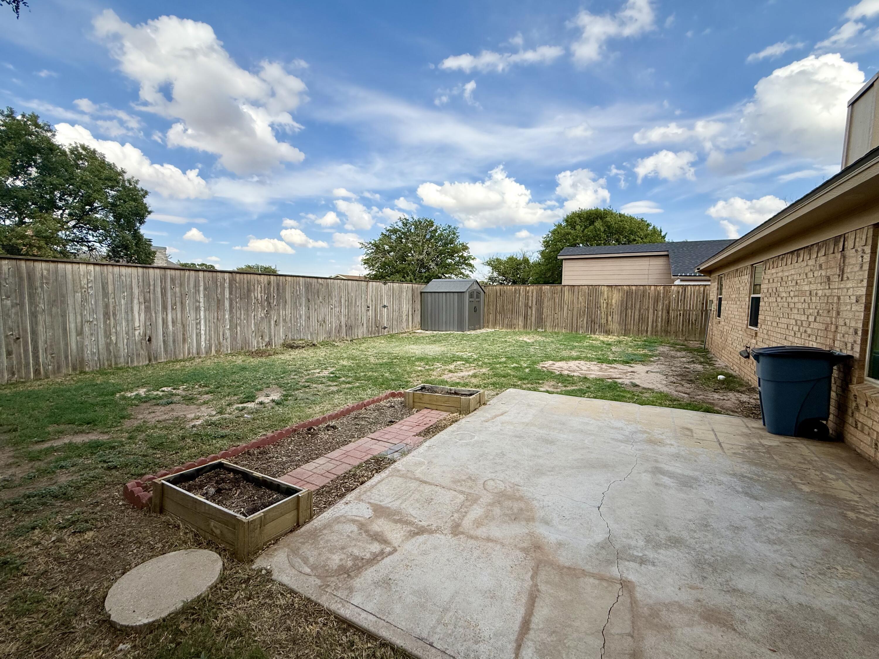 Fenced backyard with a concrete patio, raised garden beds, and a small storage shed.