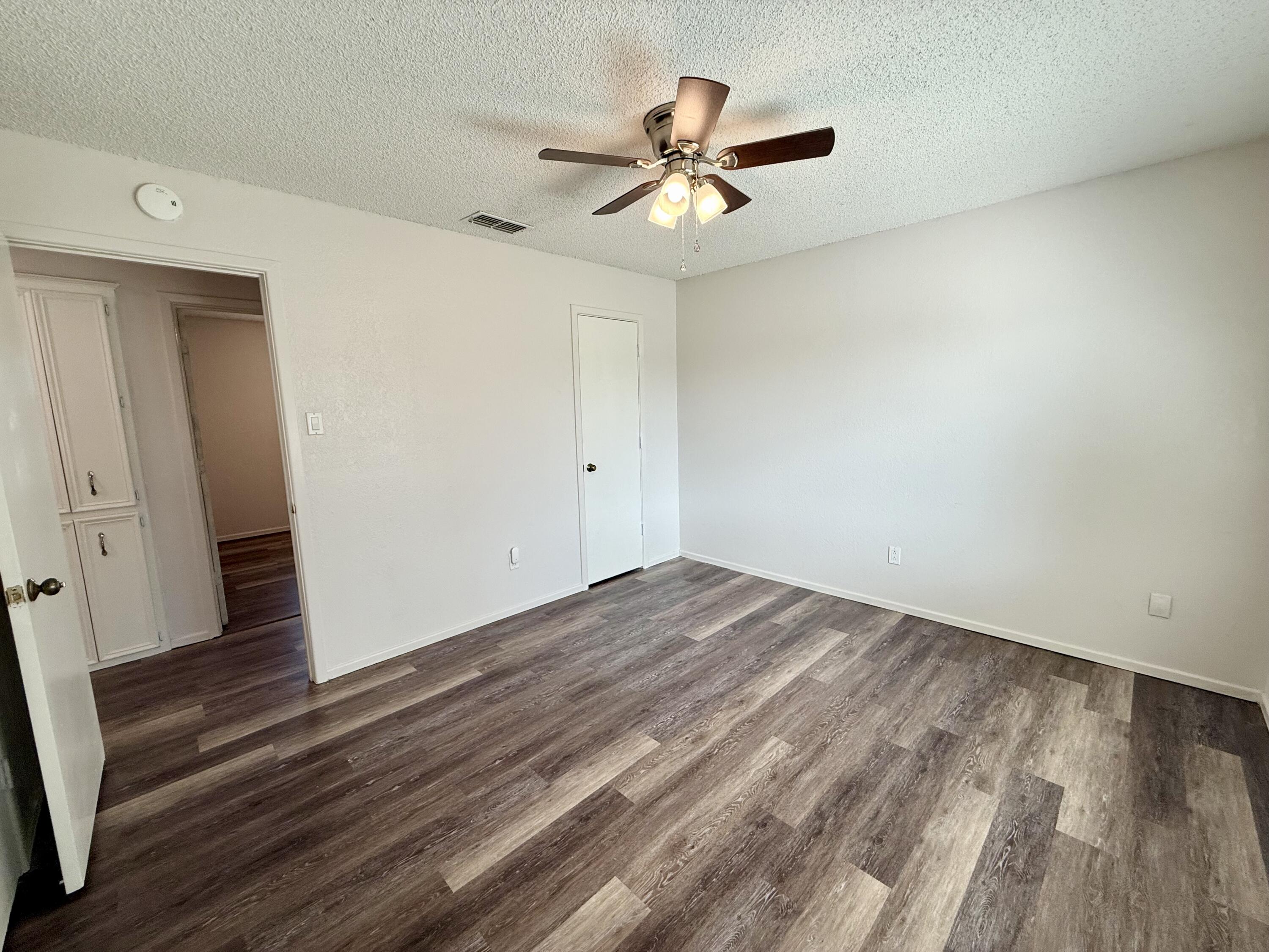 Vacant room with gray-brown plank flooring, white walls, and a lit ceiling fan.