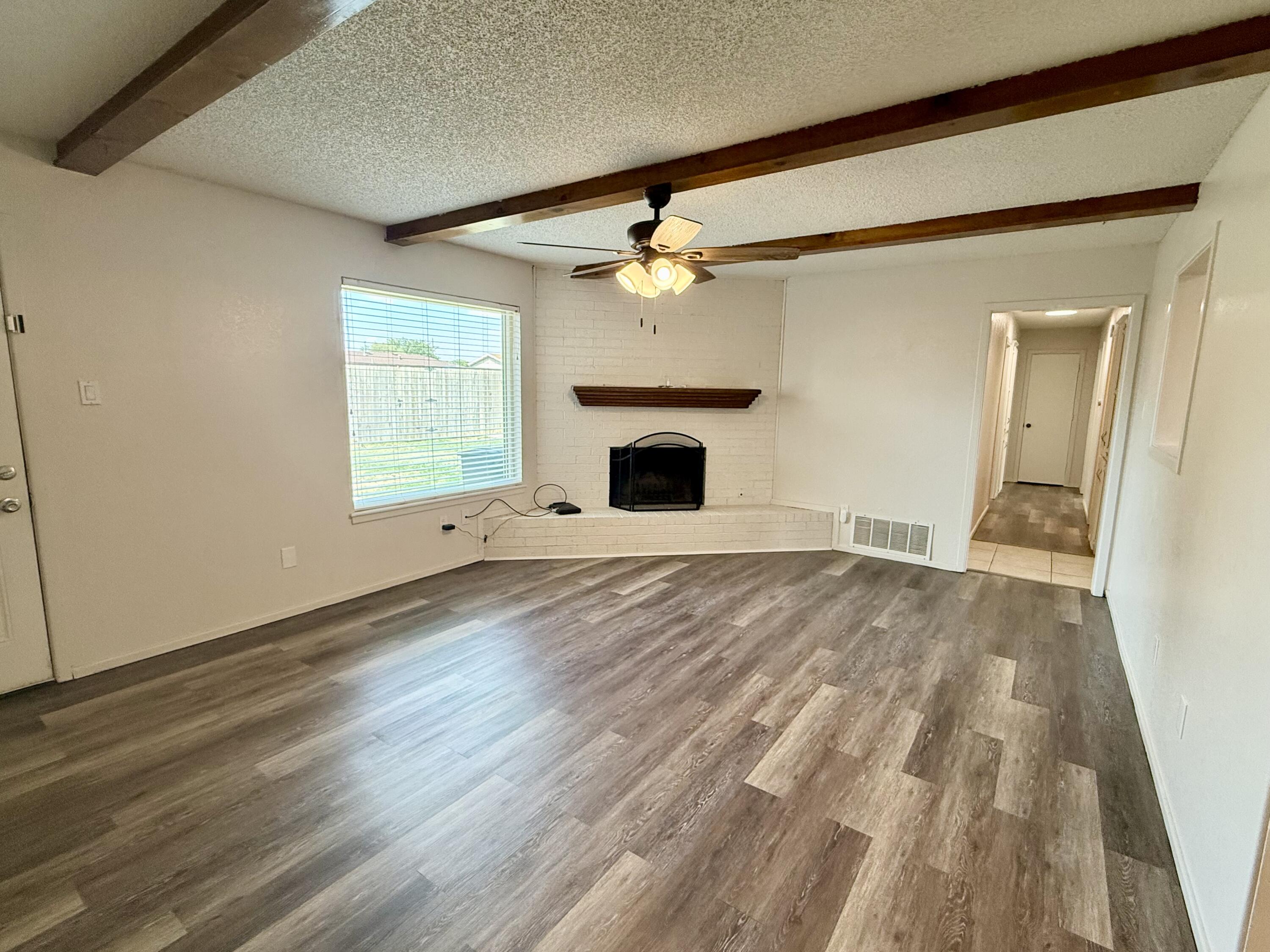 Unfurnished living room with a white brick fireplace, exposed ceiling beams, and gray-brown plank flooring.