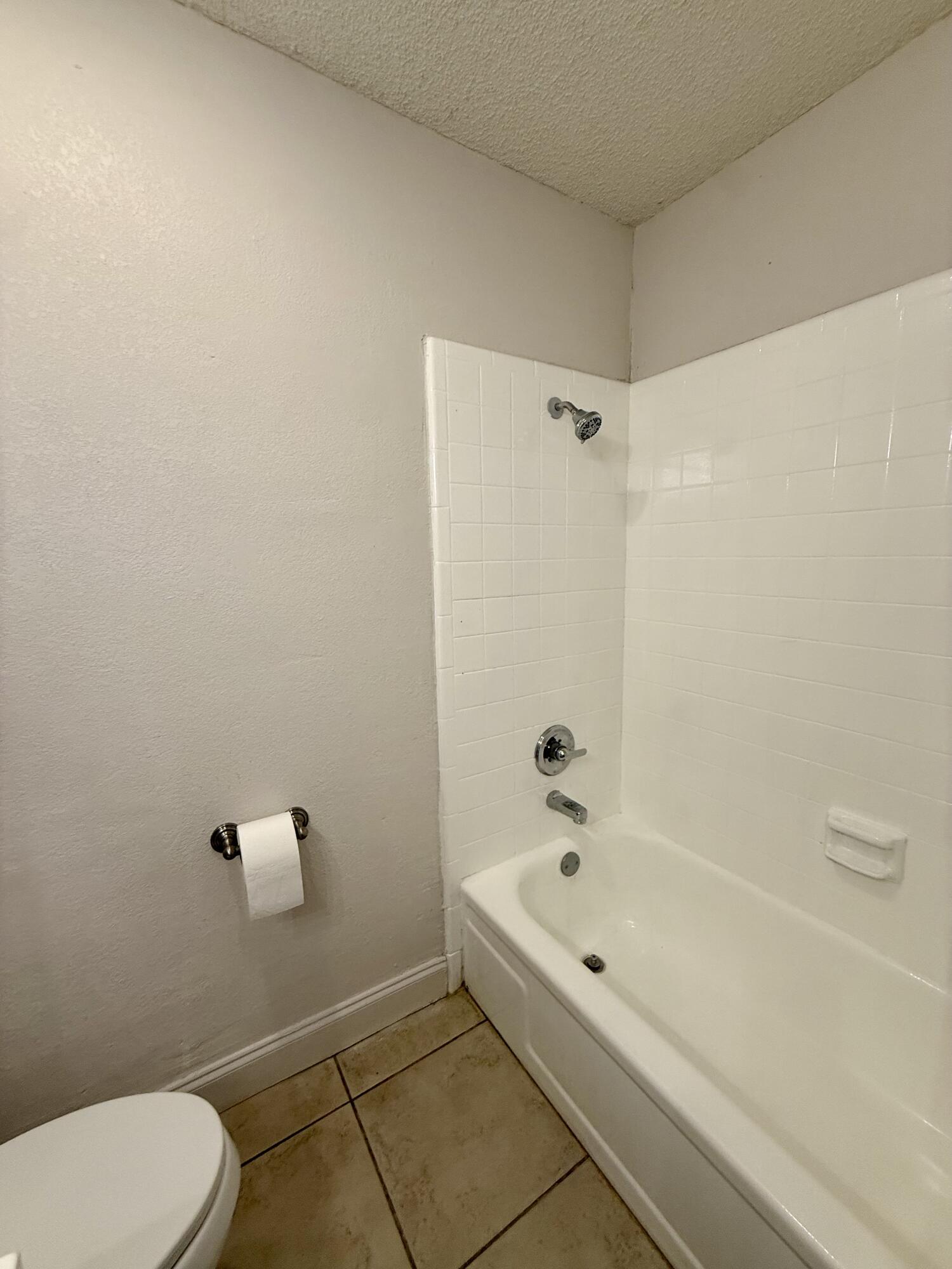 Bathroom with a white tub, tiled shower surround, chrome fixtures, and tan tile floor.