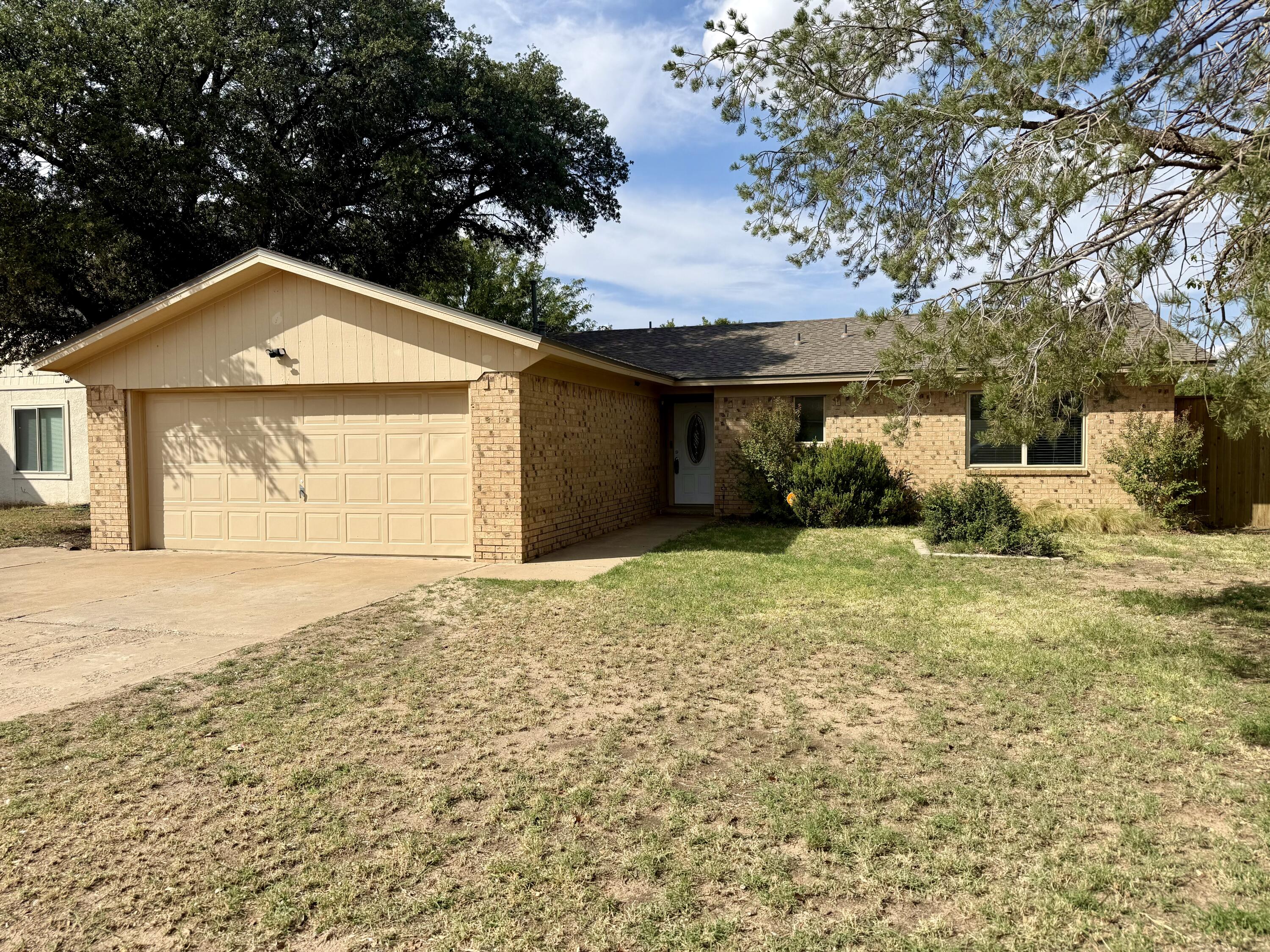 Single-story brick house with a two-car garage, shaded entry, and mature trees.