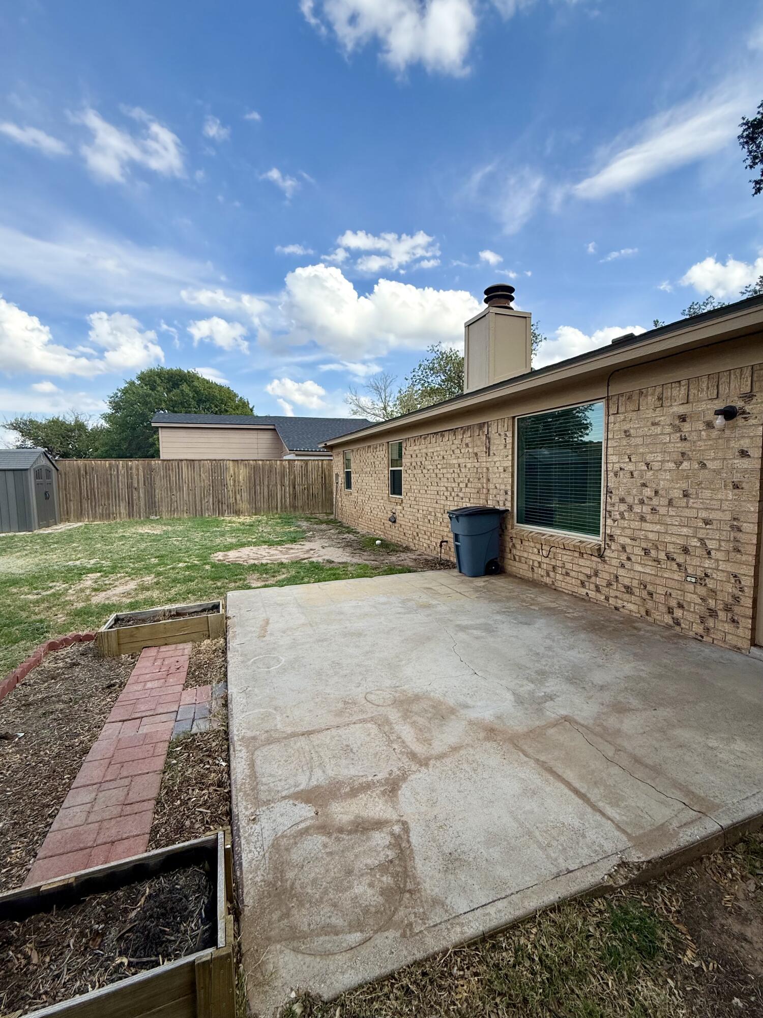 Concrete backyard patio beside a tan brick house, with raised garden beds and a wooden privacy fence.
