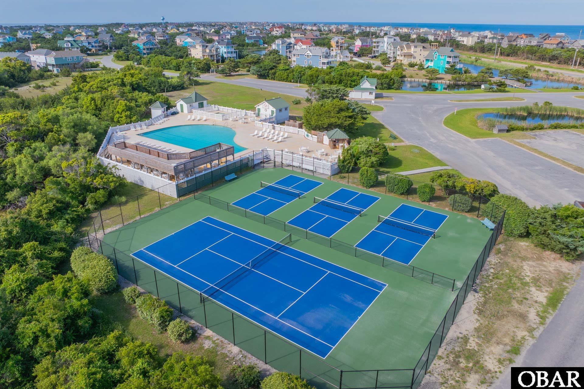 Aerial view of community tennis courts and pool, with coastal homes and ocean in the distance.