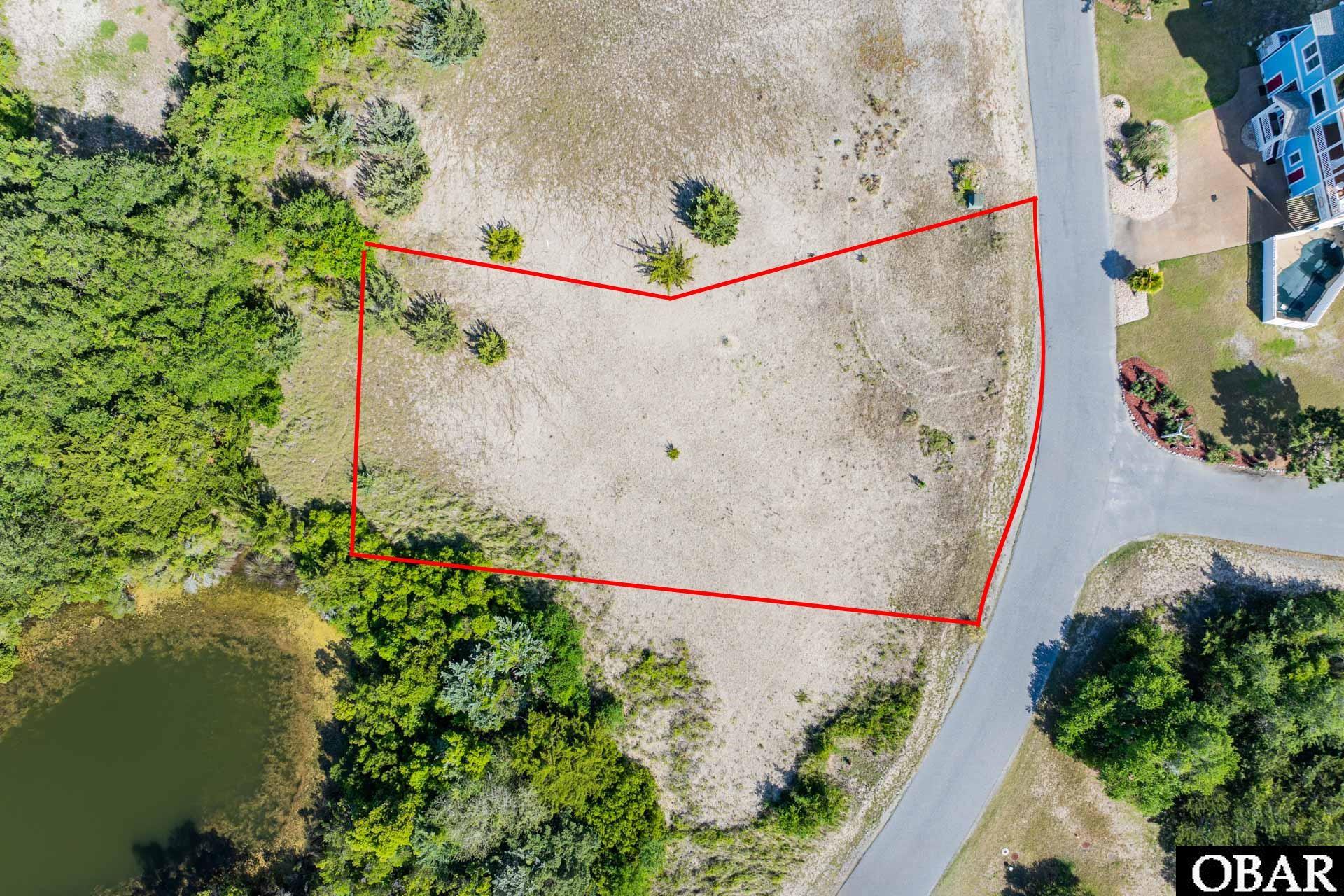 Aerial view of a vacant lot outlined in red beside a road, with trees and a pond nearby.