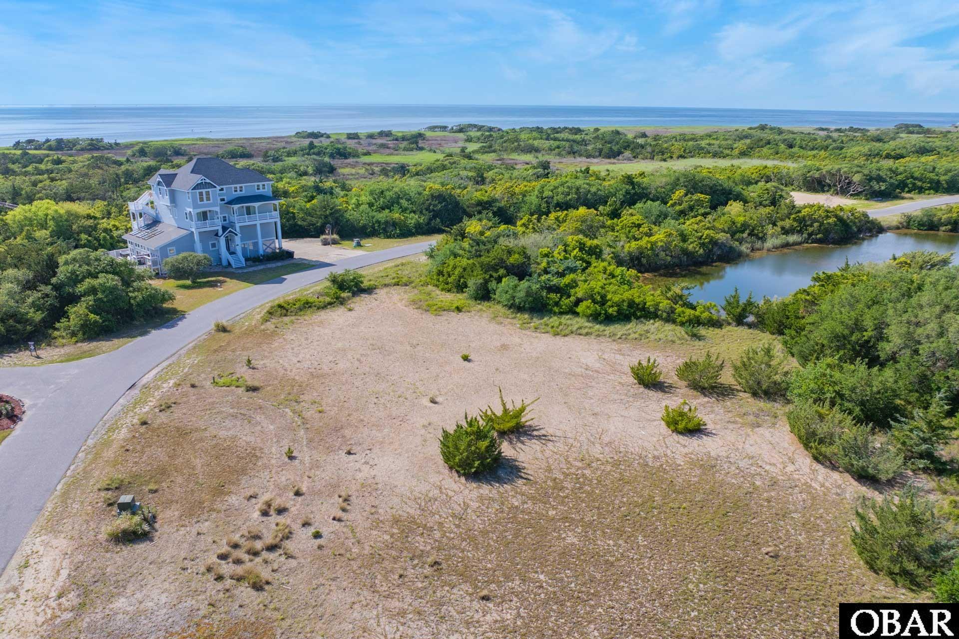 Aerial view of a vacant coastal lot with a nearby house, pond, and open water beyond.