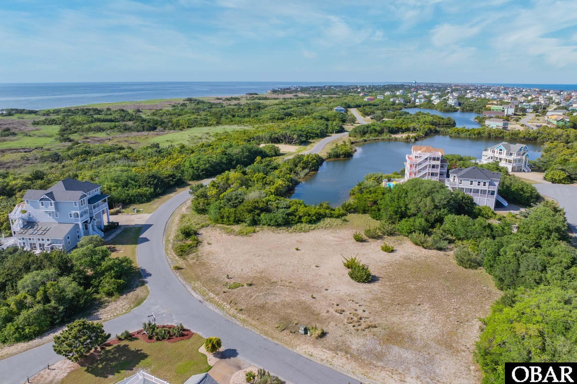 Vacant coastal lot beside a curving road, with a pond, nearby homes, and ocean beyond the marsh.
