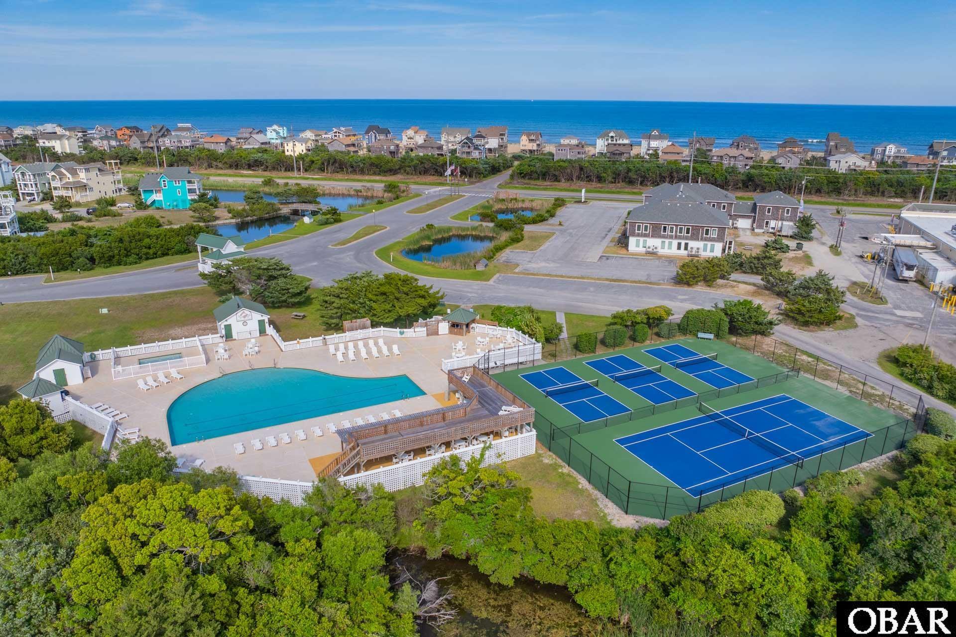 Aerial view of a coastal neighborhood with a community pool, tennis courts, and ocean beyond.