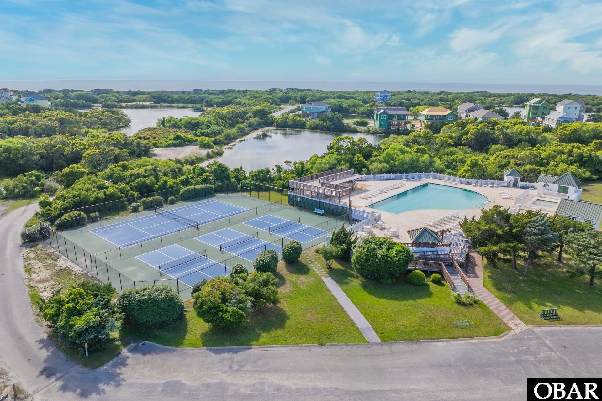 Aerial view of community tennis courts and swimming pool beside wooded wetlands and coastal water.