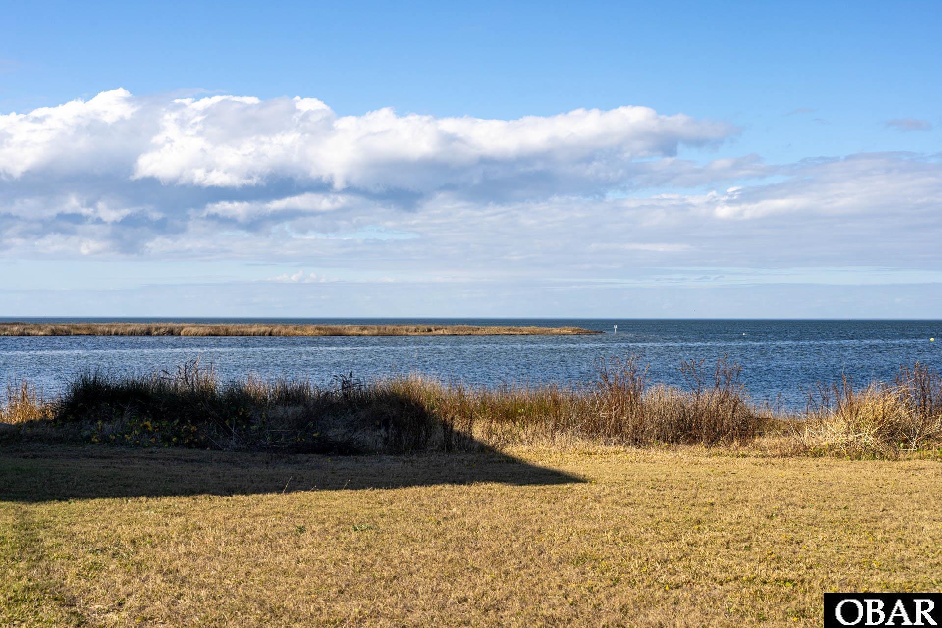 Waterfront lot with grassy foreground, calm water, and a low marshy shoreline beneath a cloud-filled sky.