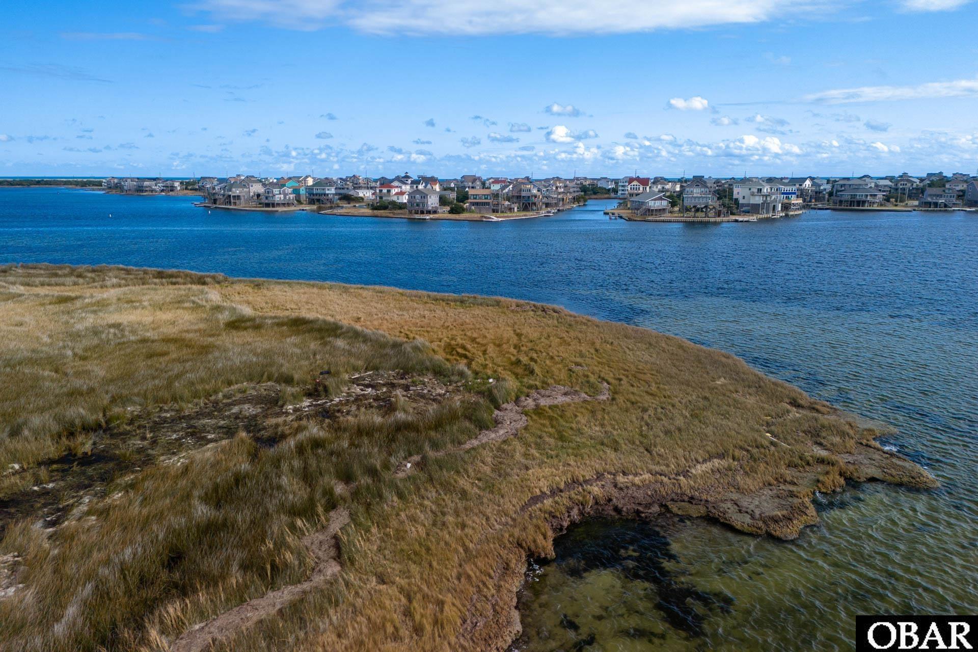 Marshy waterfront lot beside a blue inlet, with coastal homes across the water.