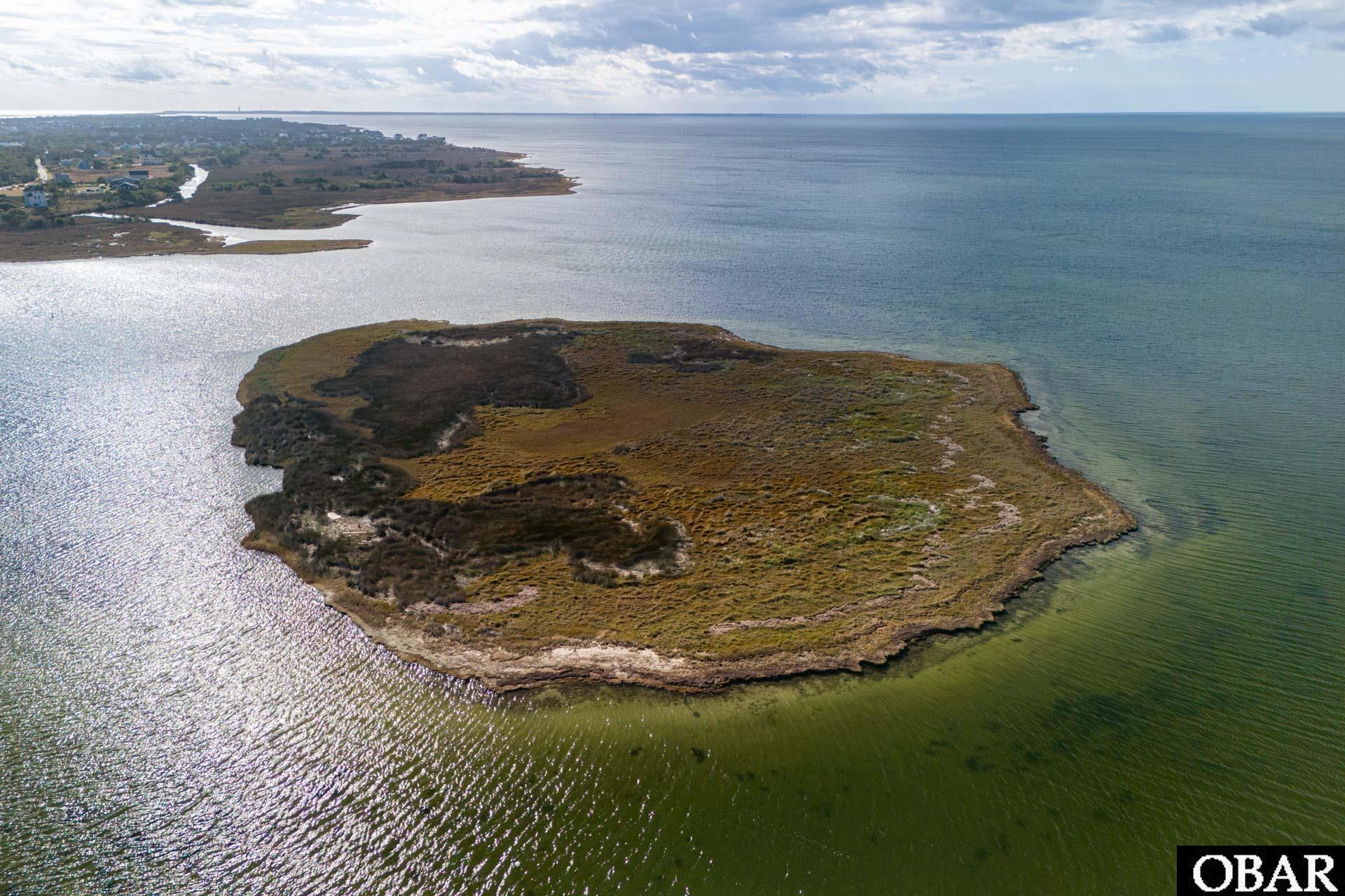 Aerial view of a marsh-covered island lot surrounded by water, with distant homes along the shoreline.