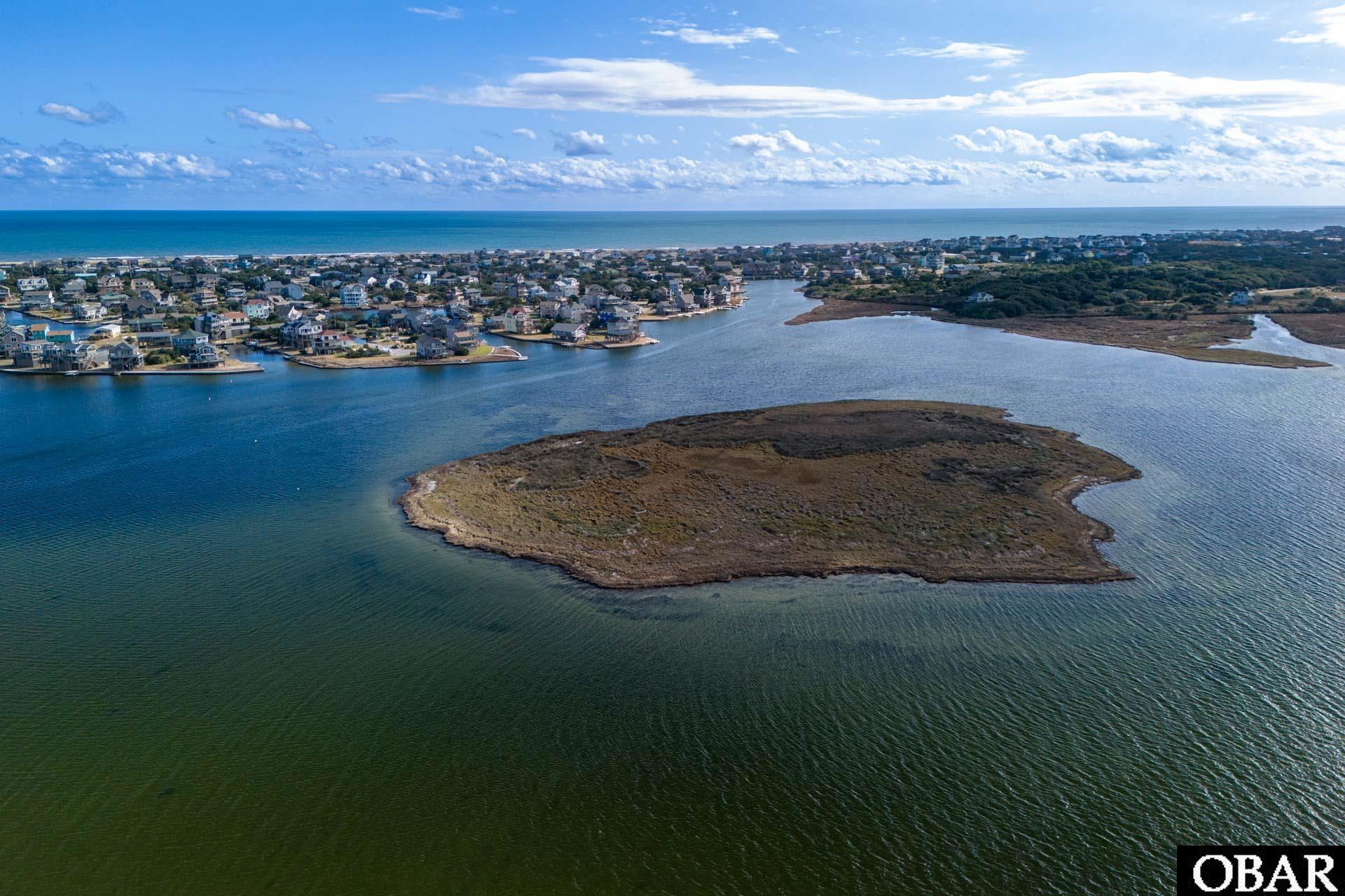 Aerial view of a marshy island lot surrounded by water, with coastal homes and ocean beyond.
