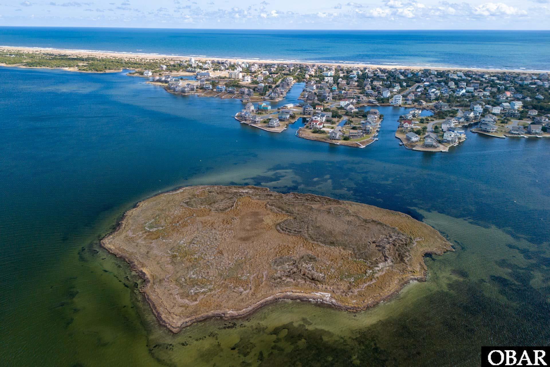 Undeveloped island lot surrounded by water, with a waterfront neighborhood and ocean beach beyond.