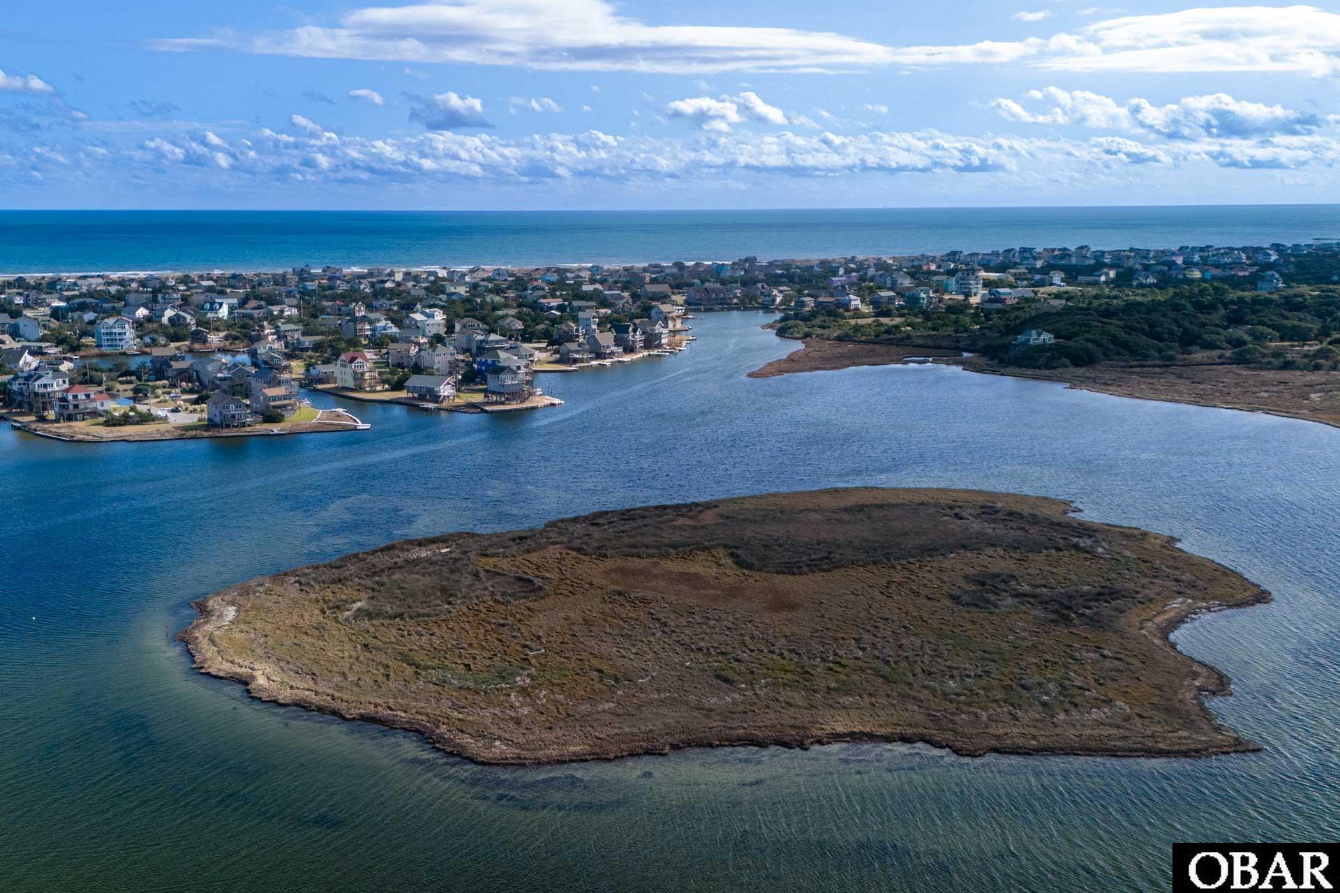Aerial view of undeveloped marsh island surrounded by water, with coastal homes and ocean beyond.
