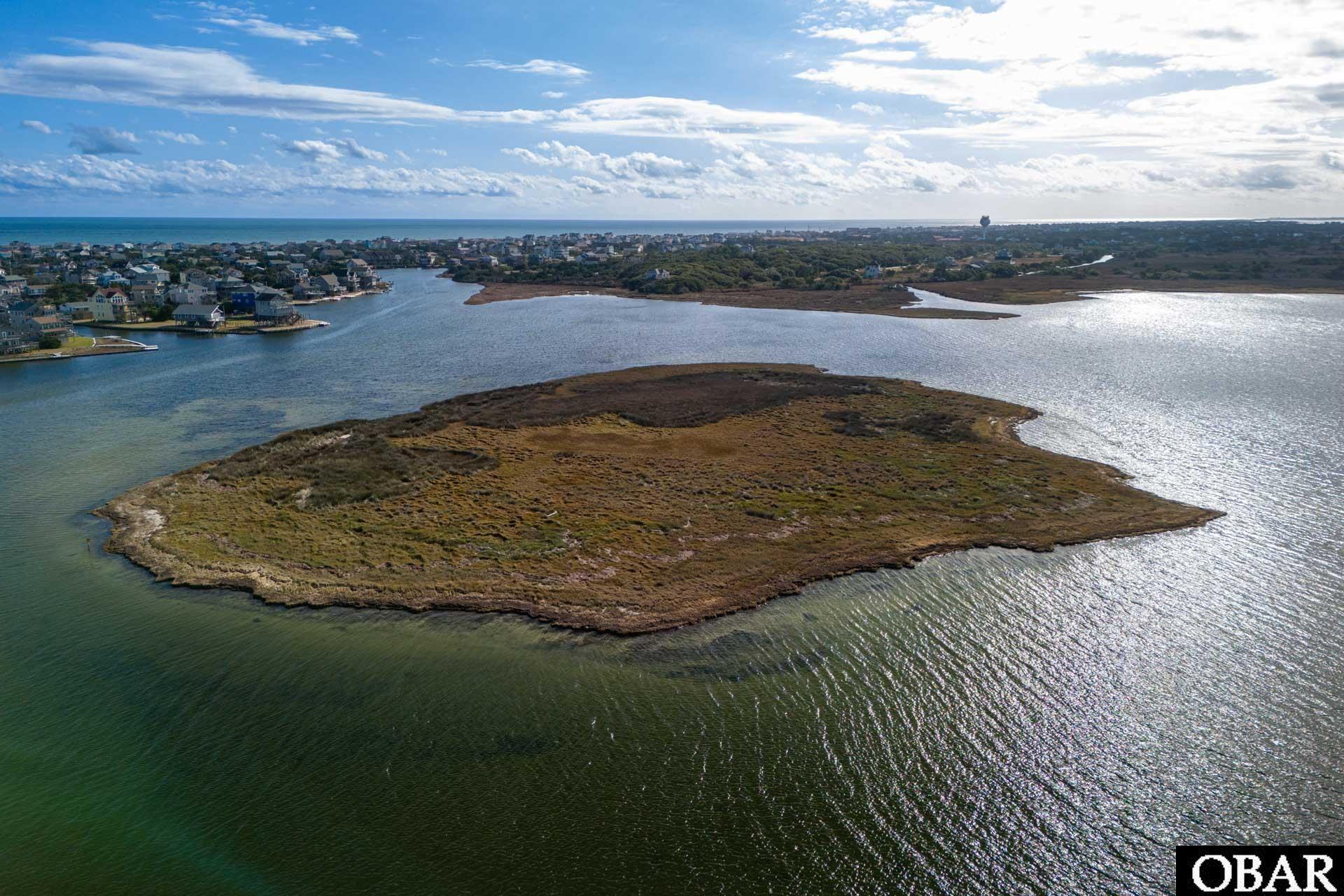 Aerial view of a marshy island lot surrounded by water, with coastal homes and ocean in the distance.
