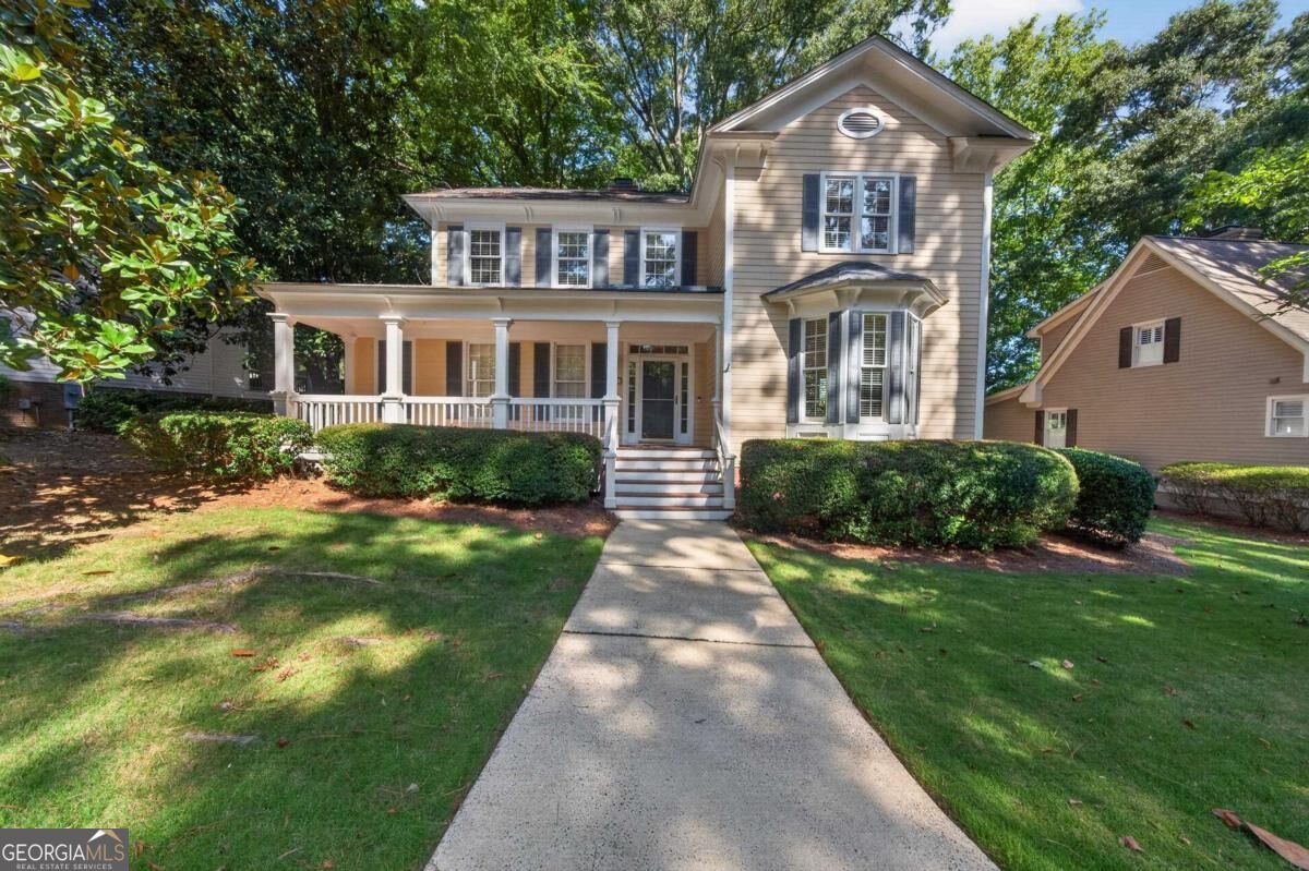 Two-story beige house with a columned porch, bay window, and walkway through a shaded lawn.