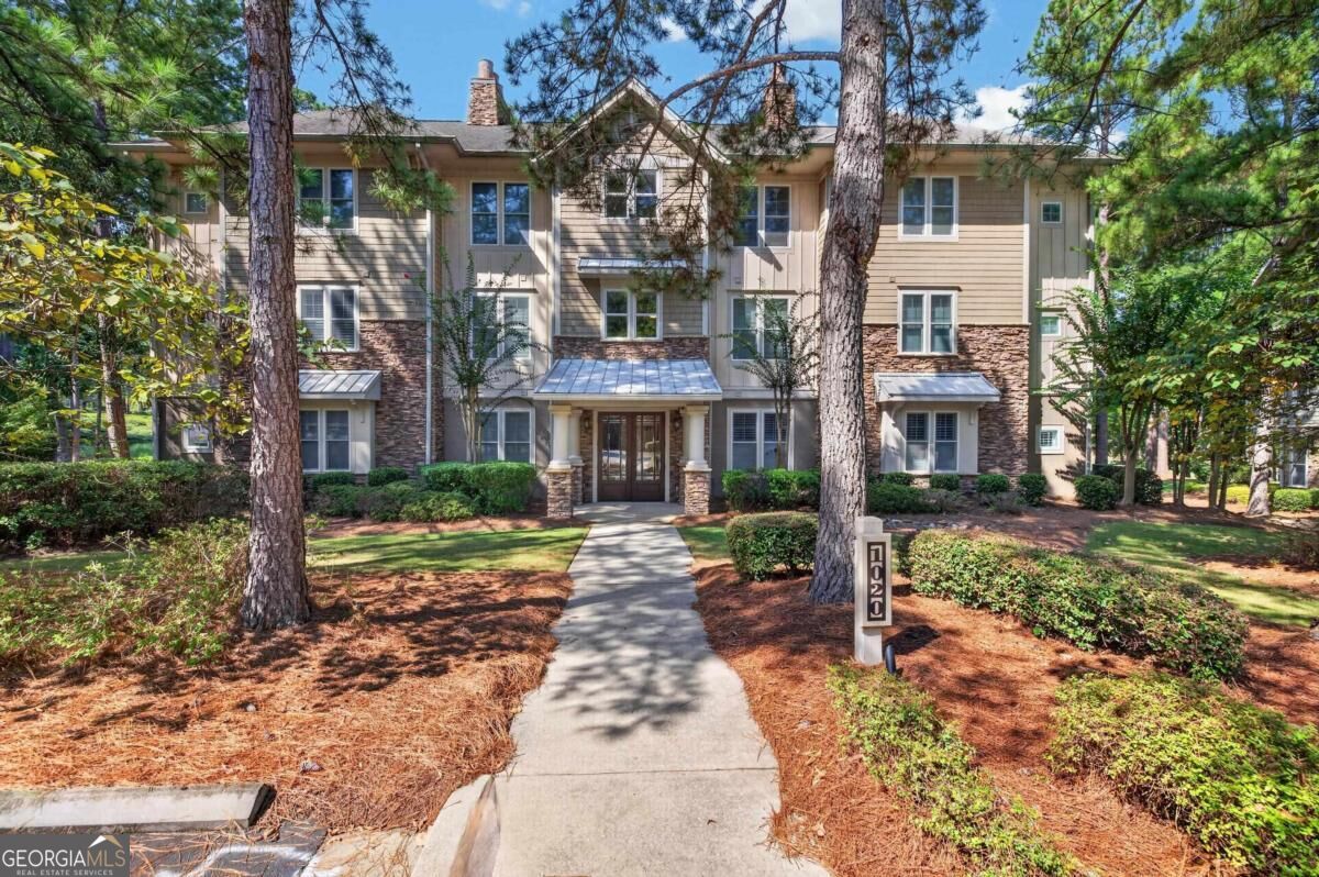 Three-story beige residence with stone veneer, a covered entrance, and a landscaped walkway beneath tall pines.