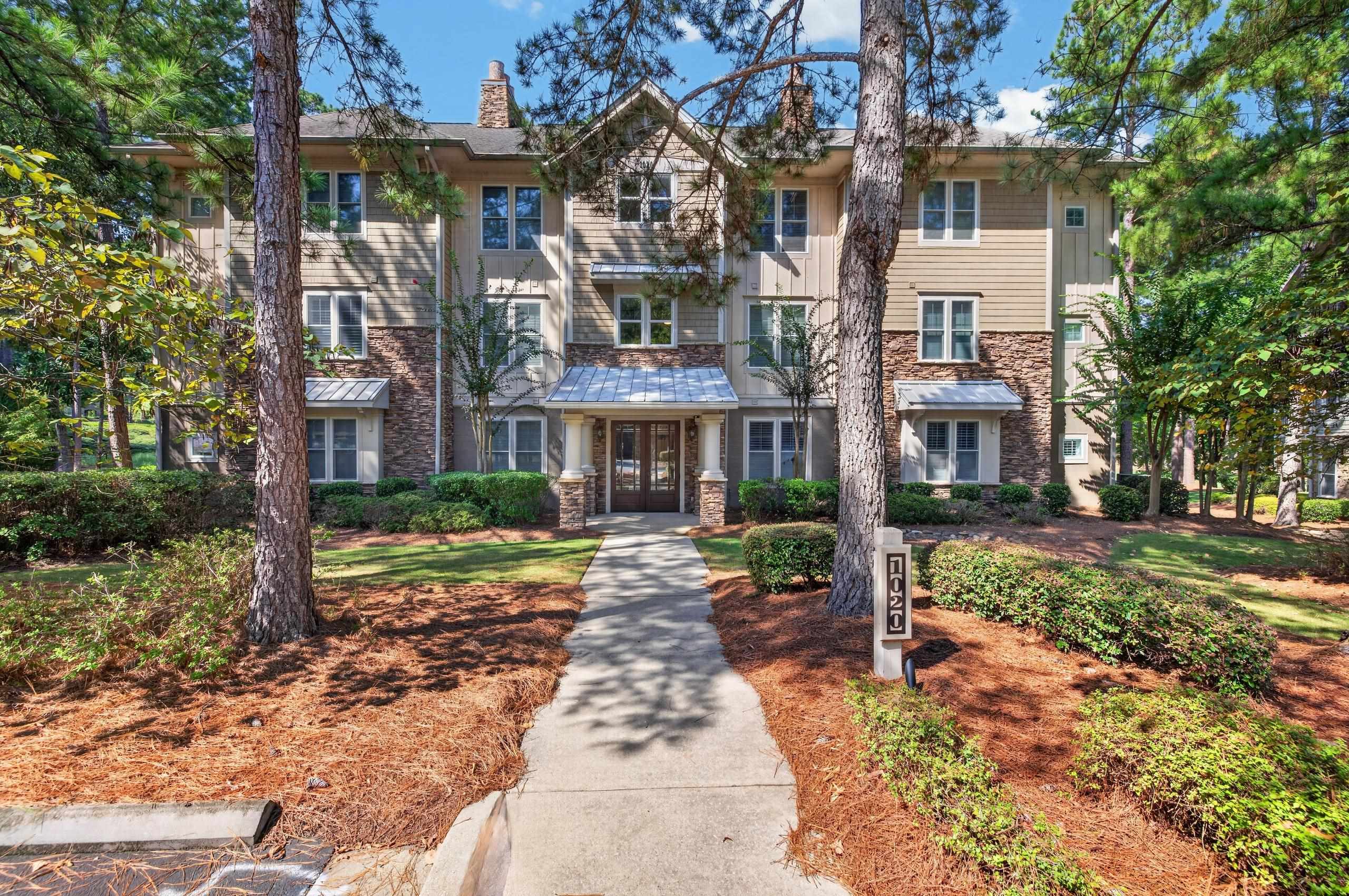 Residential building with stone accents and a covered entrance, approached by a landscaped walkway beneath pine trees.