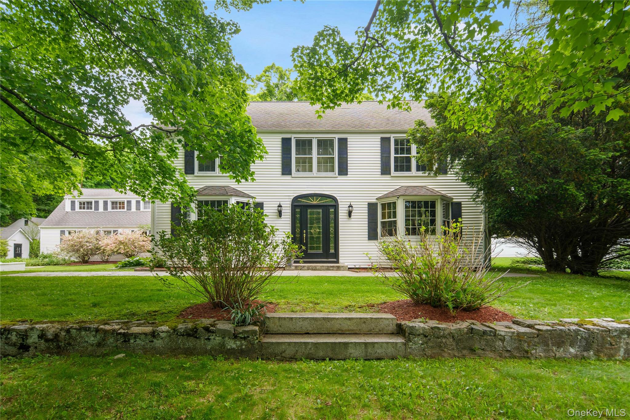 White two-story house with dark shutters, bay windows, and a landscaped lawn behind a stone wall.