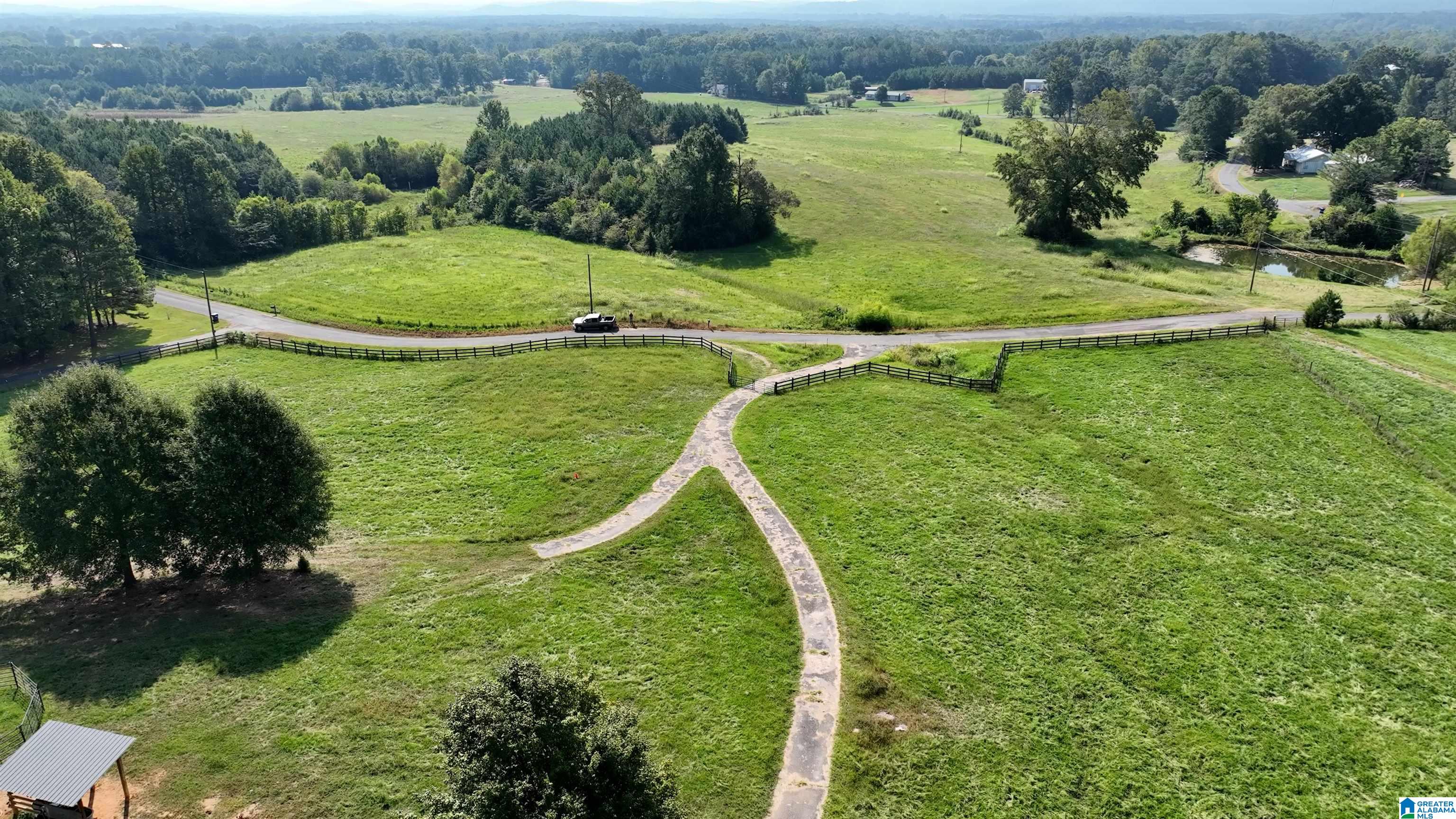 Aerial view of green rural acreage with a forked path, fenced road, scattered trees, and distant wooded hills.