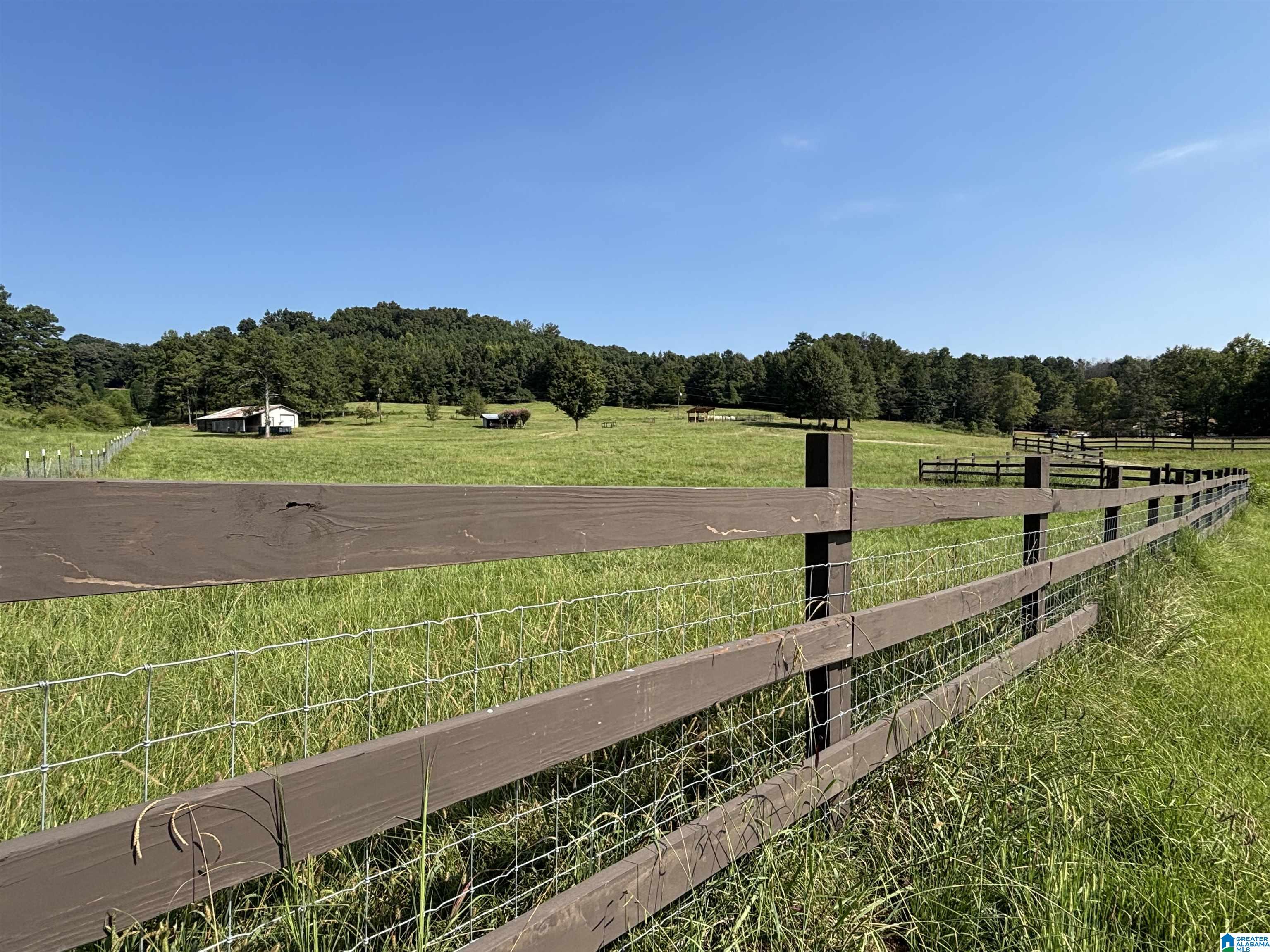 Fenced green pasture with tall grass, small outbuildings, and wooded hills beyond.