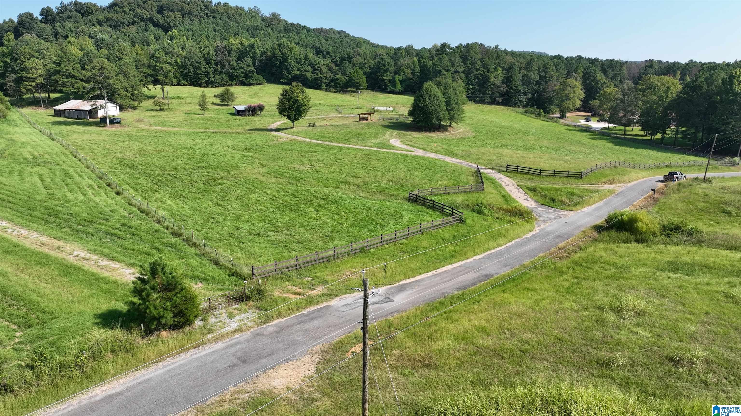 Aerial view of fenced green pasture with barns, wooded hills, and a road.