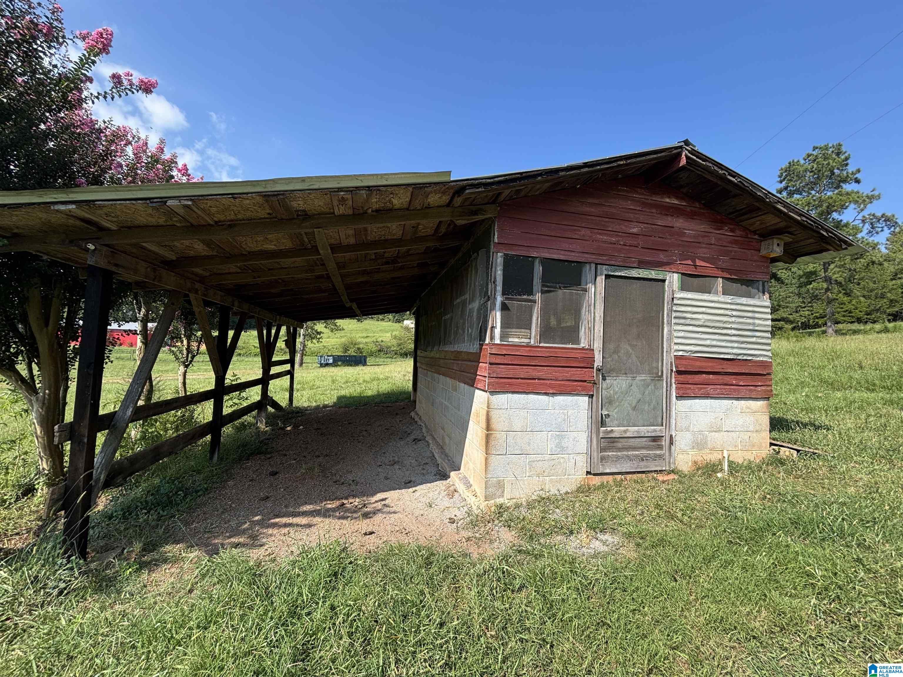 Red-sided outbuilding with screened windows and an attached open-sided covered shelter in a grassy yard.