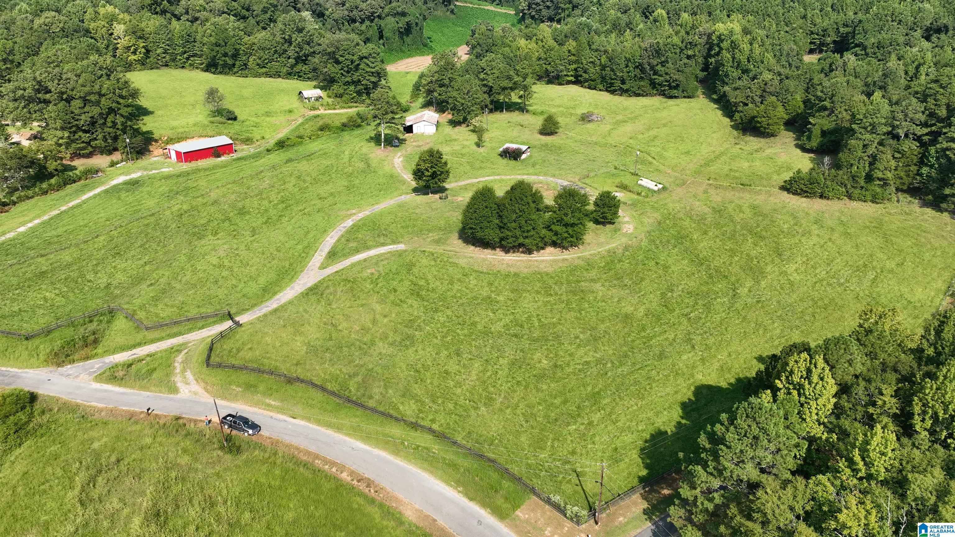 Aerial view of a green pasture with several barns, a winding driveway, and wooded borders.