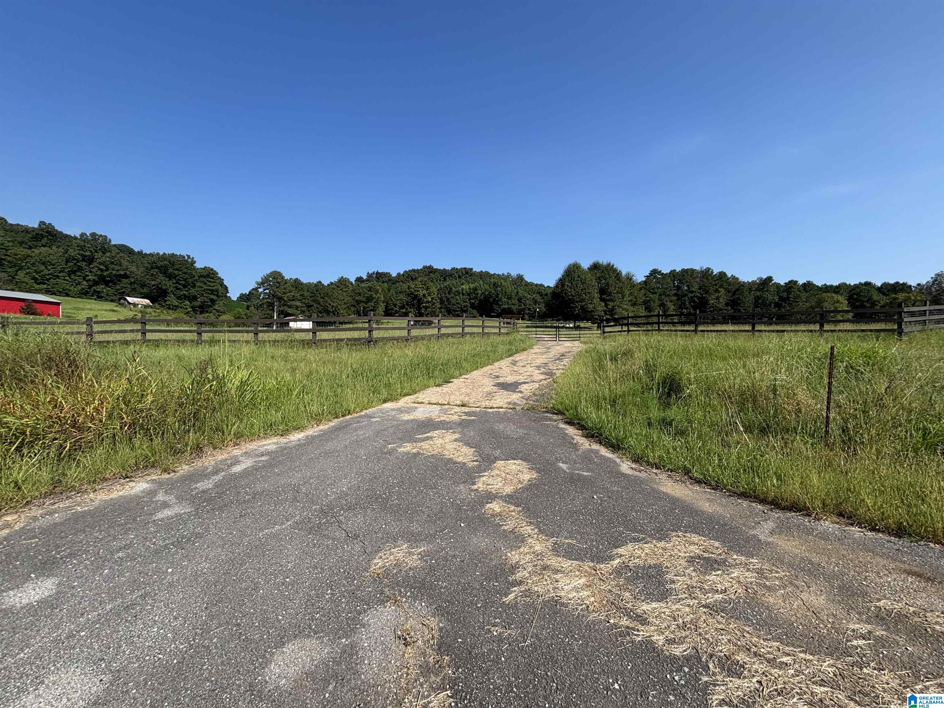 Paved drive crosses fenced green pasture toward a gate, with a red barn and wooded hills beyond.