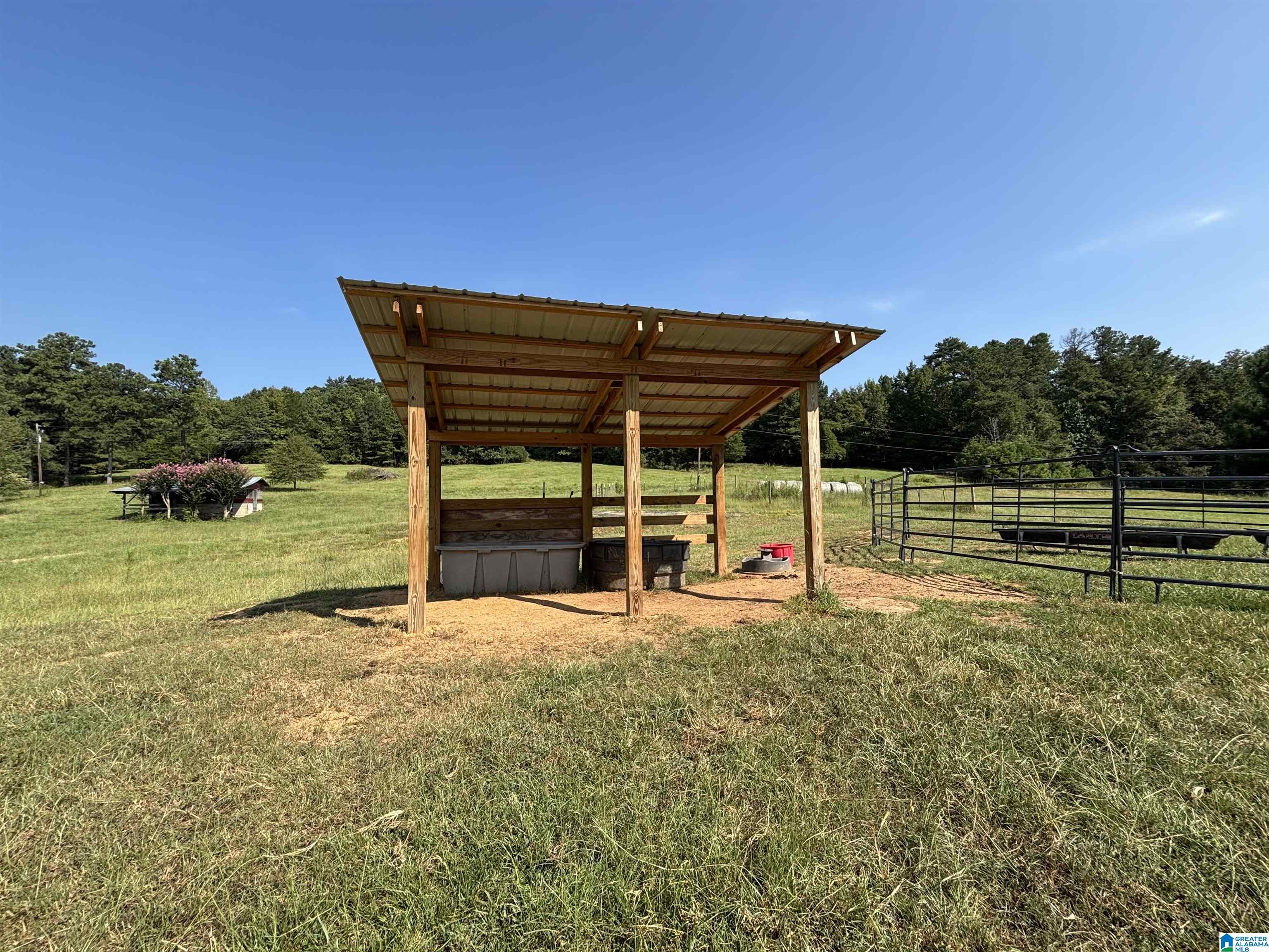Open-sided wooden livestock shelter in a grassy pasture beside metal fencing.