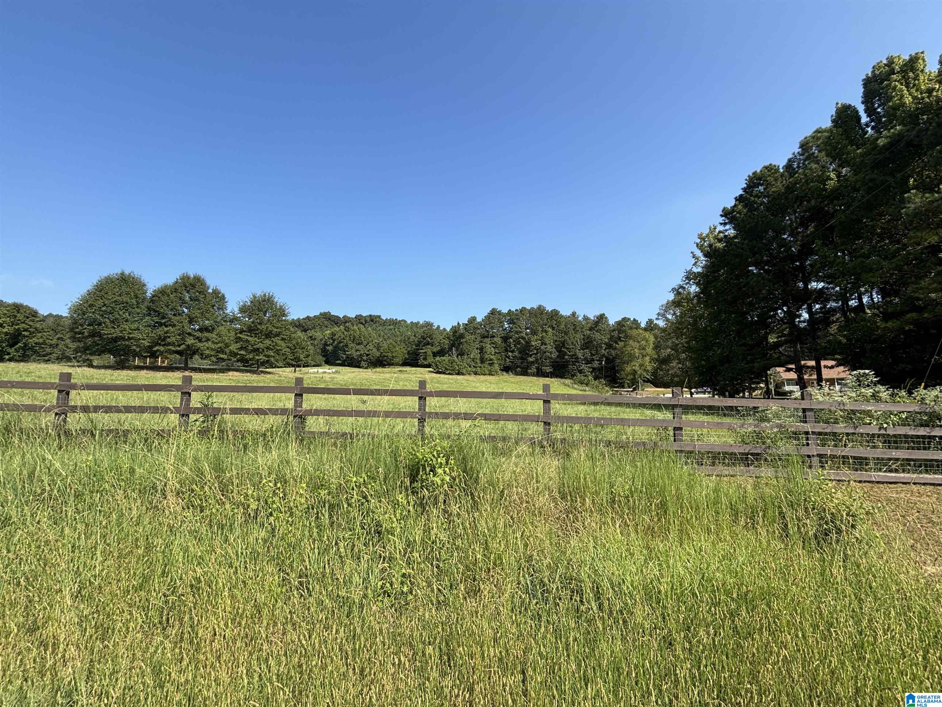 Grassy field behind a dark wooden fence, with trees and part of a house in the distance.