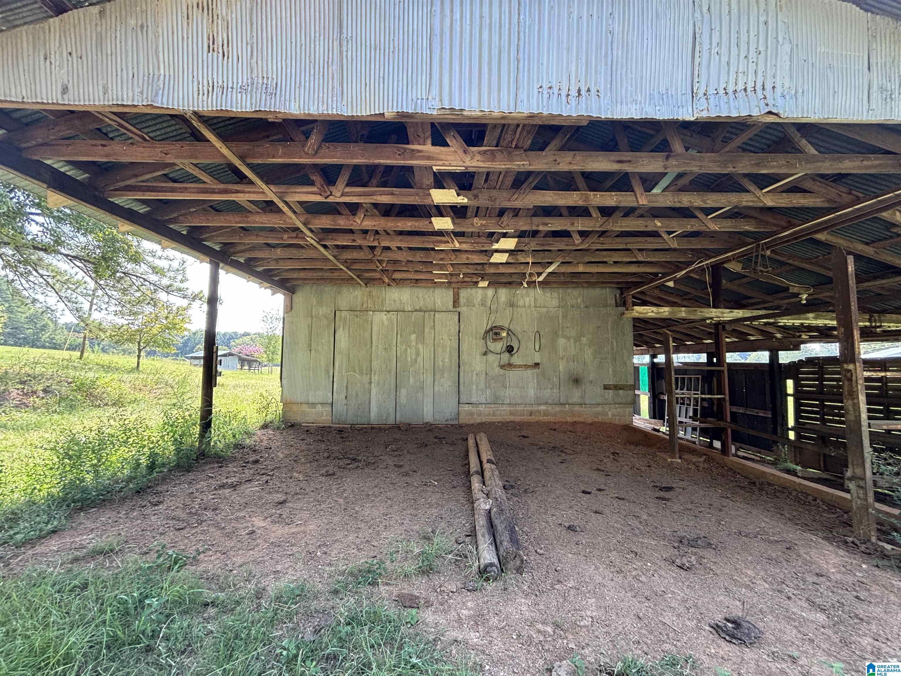 Open-sided barn with exposed wooden roof trusses, corrugated metal panels, and a dirt floor.