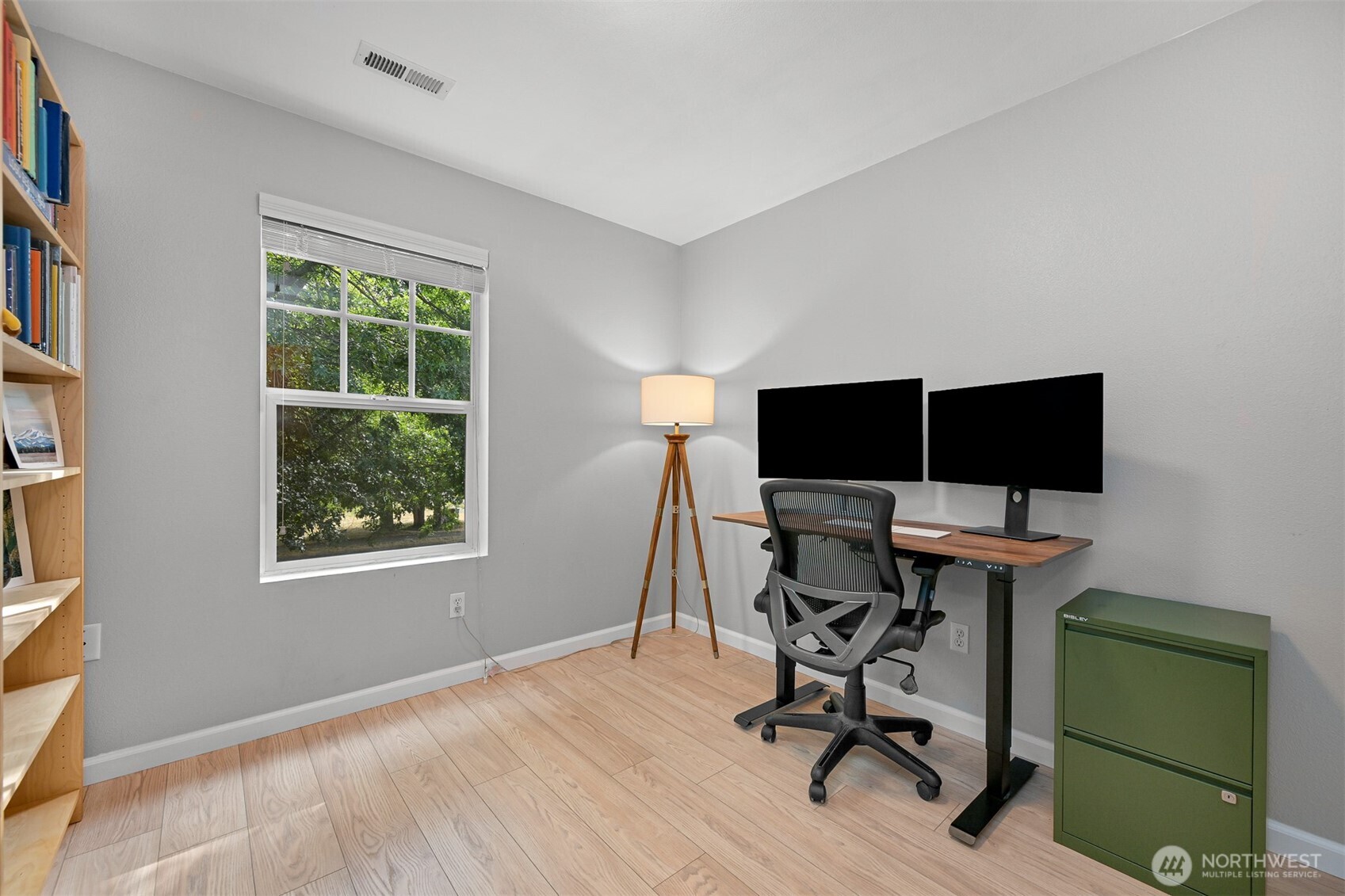 Home office with a dual-monitor desk, green filing cabinet, and window overlooking trees.