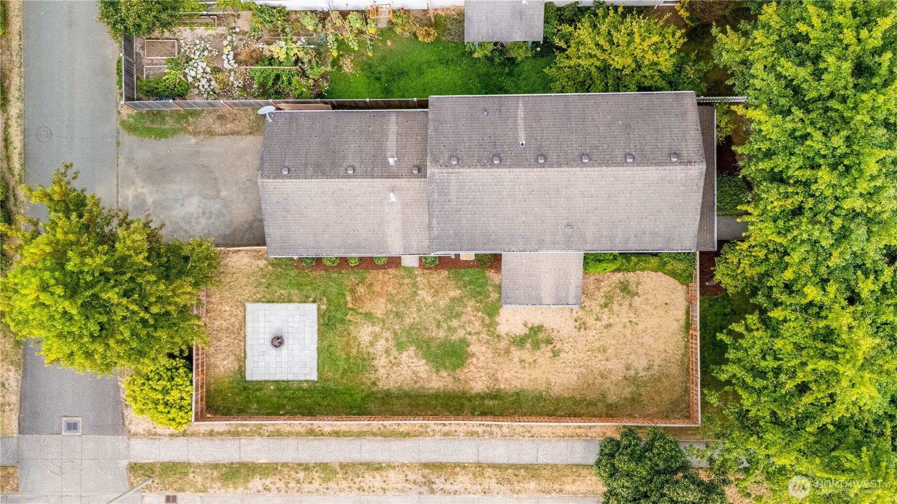 Aerial view of a home with a fenced, patchy lawn, patio, fire pit, and surrounding trees.