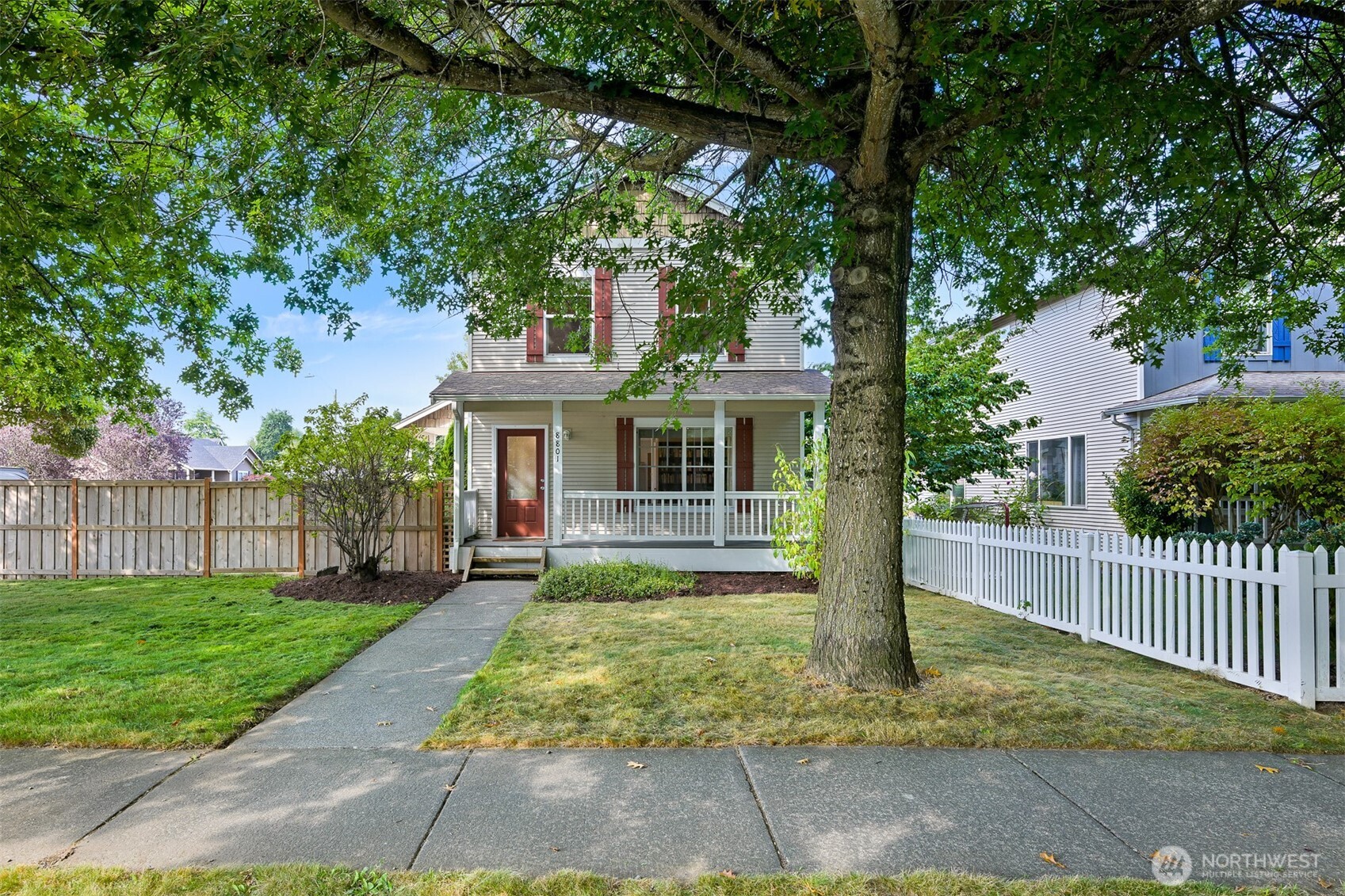 Two-story house with a covered porch, red shutters, and a large shade tree in the front yard.