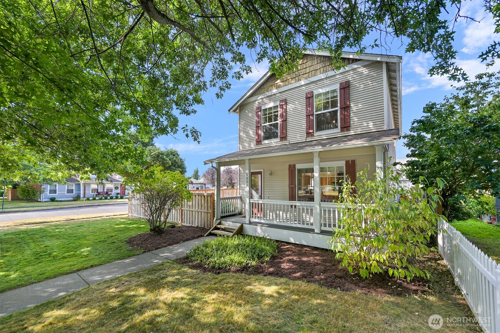 Two-story house with red shutters, a covered porch, and a leafy fenced yard.