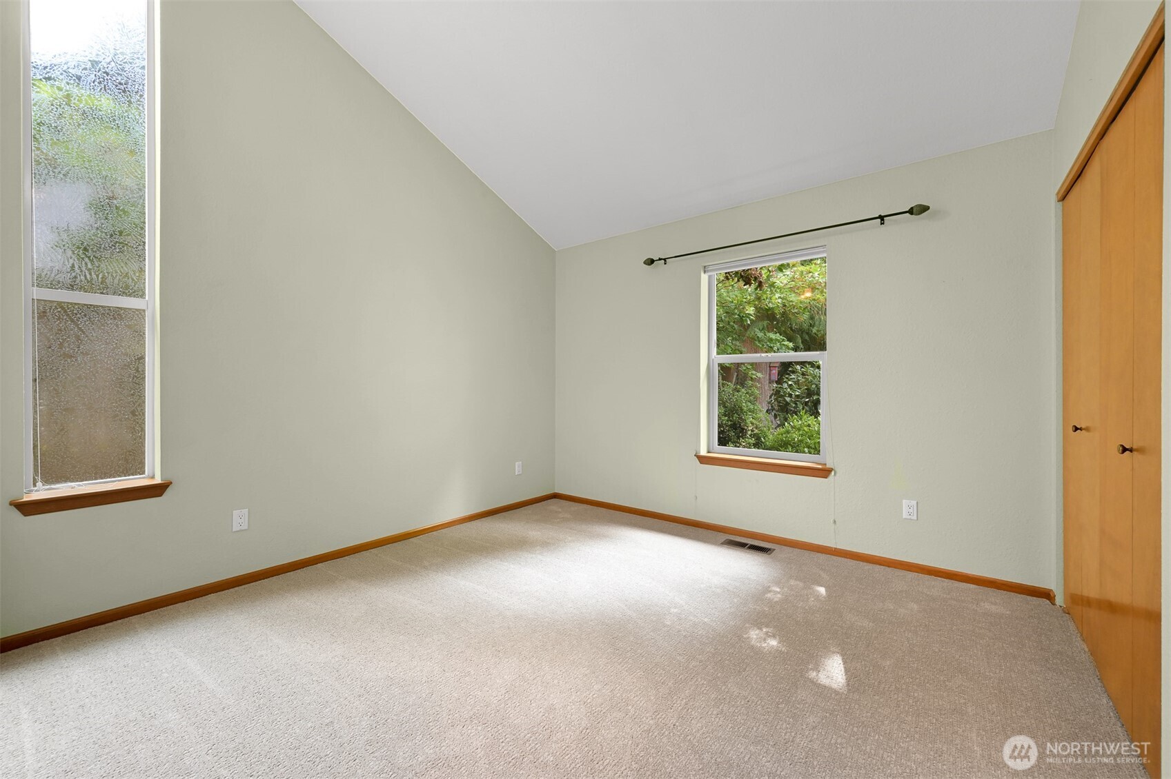 Empty bedroom with beige carpet, vaulted ceiling, two windows, and a built-in wooden closet.