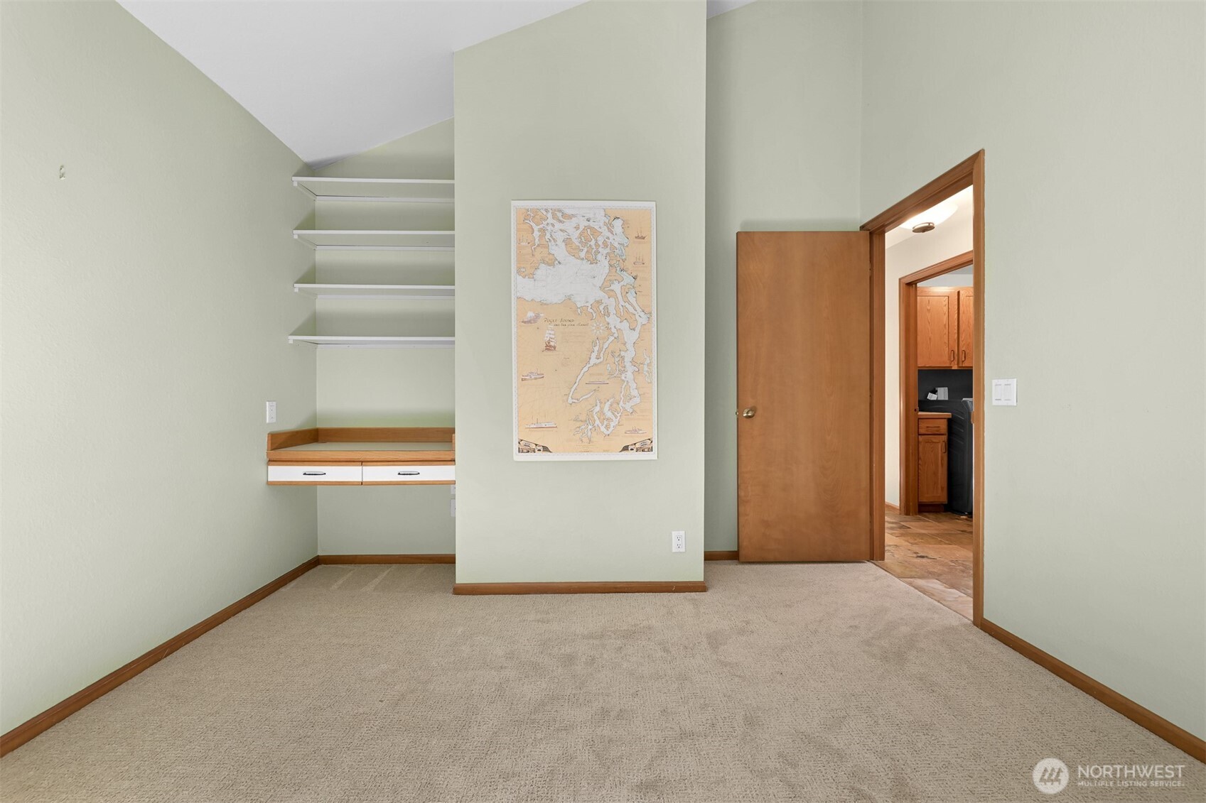Carpeted room with built-in desk and shelves, vaulted ceiling, and doorway to a tiled area.