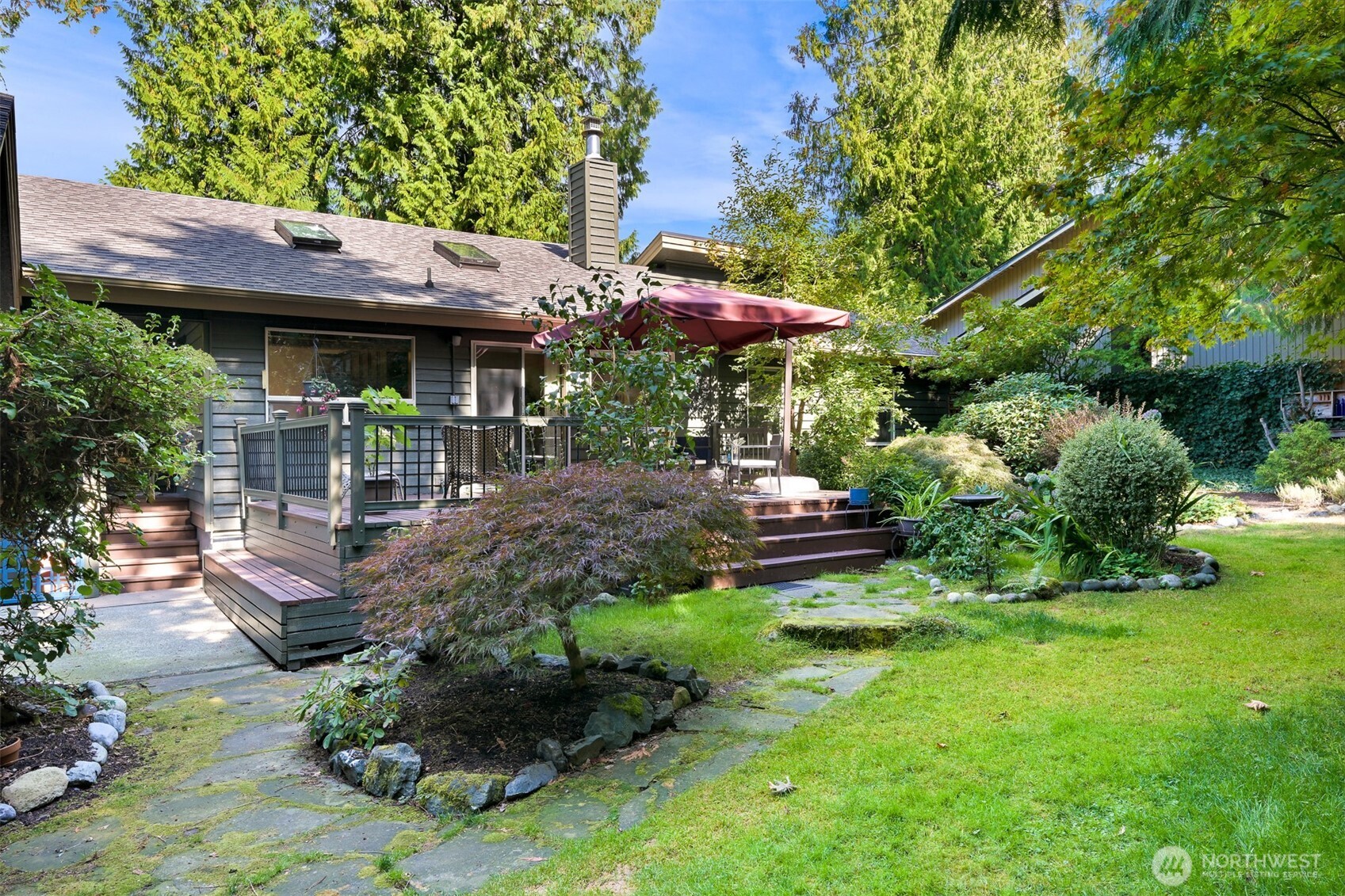 House’s raised deck with a burgundy umbrella, surrounded by a landscaped garden and stone path.